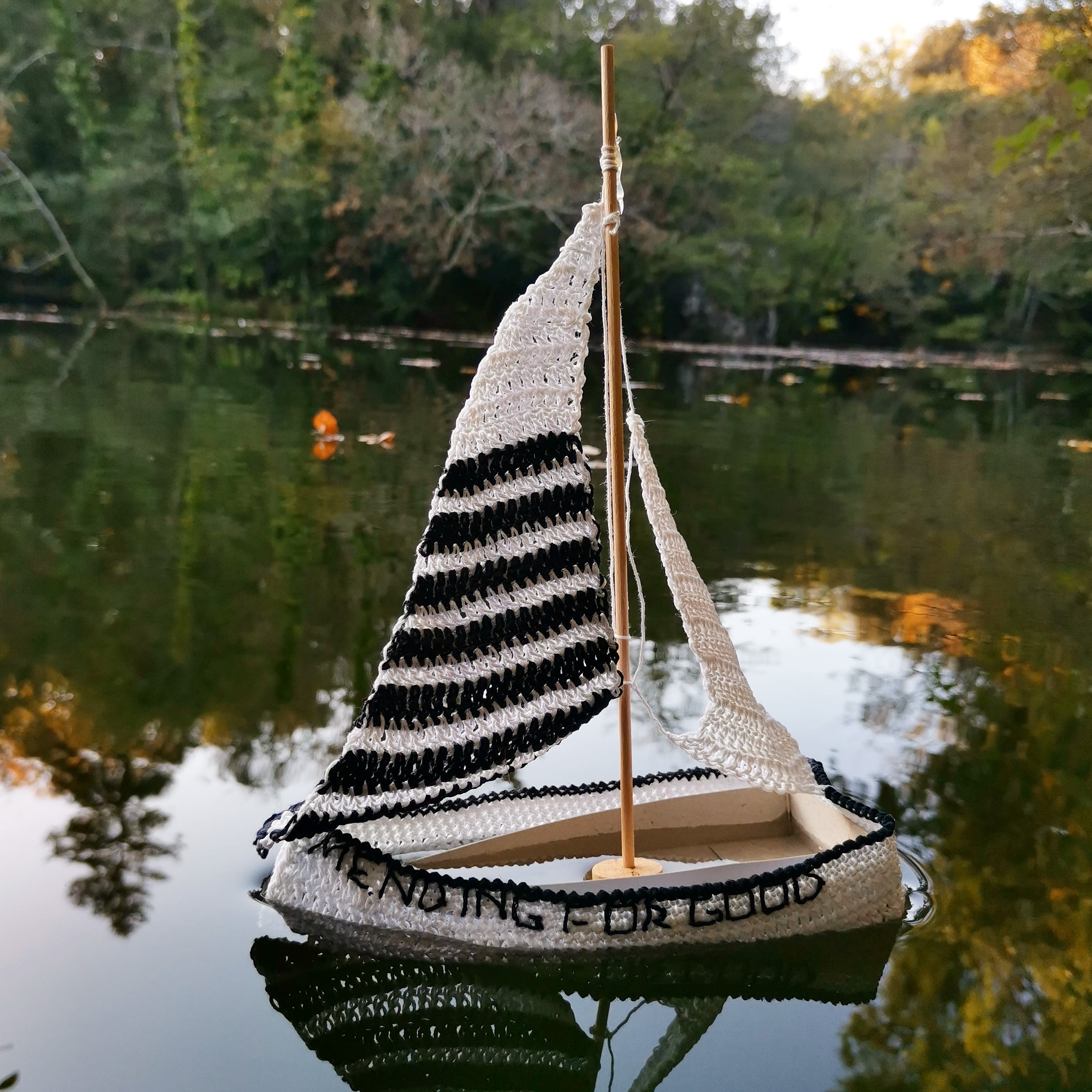 wooden model sized boat covered in a crochet monochrome design, with crocheted sails, floating on a body of water