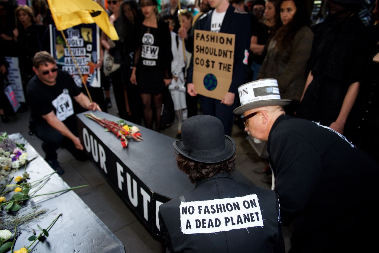 Three people hold a black coffin that says 'our future' on it. On the back on one of their jackets it says 'no fashion on a dead planet'. A person in the crowd holds a sign saying 'fashion shouldn't cost the earth'.