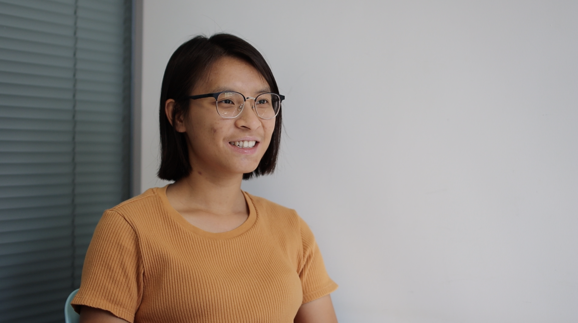 a person with short black hair and clear glasses smiling in front of a grey wall