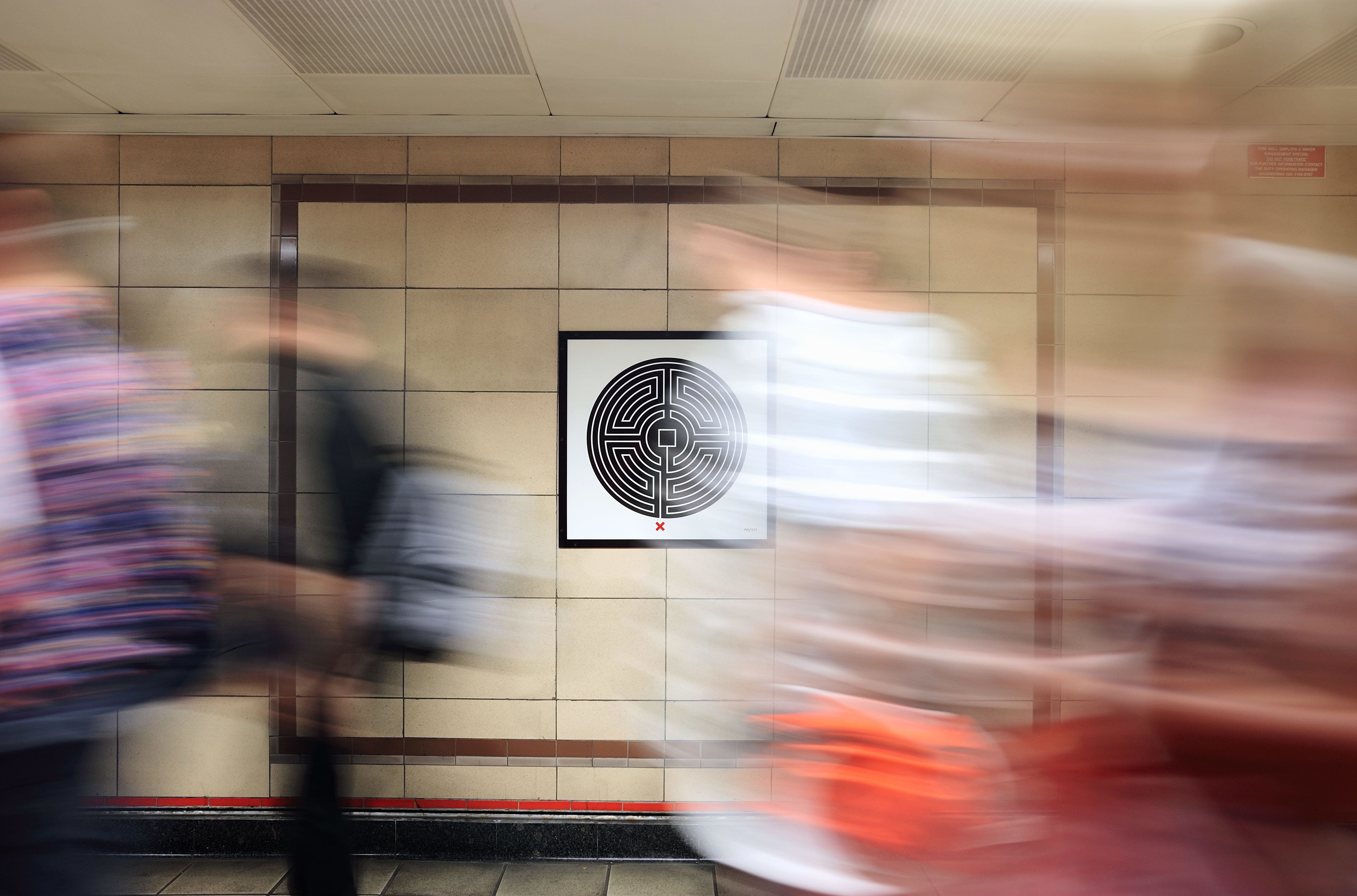 Maze artwork with a red dot at the bottom displayed on the wall of a tube station, Blurred figures are moving past. 