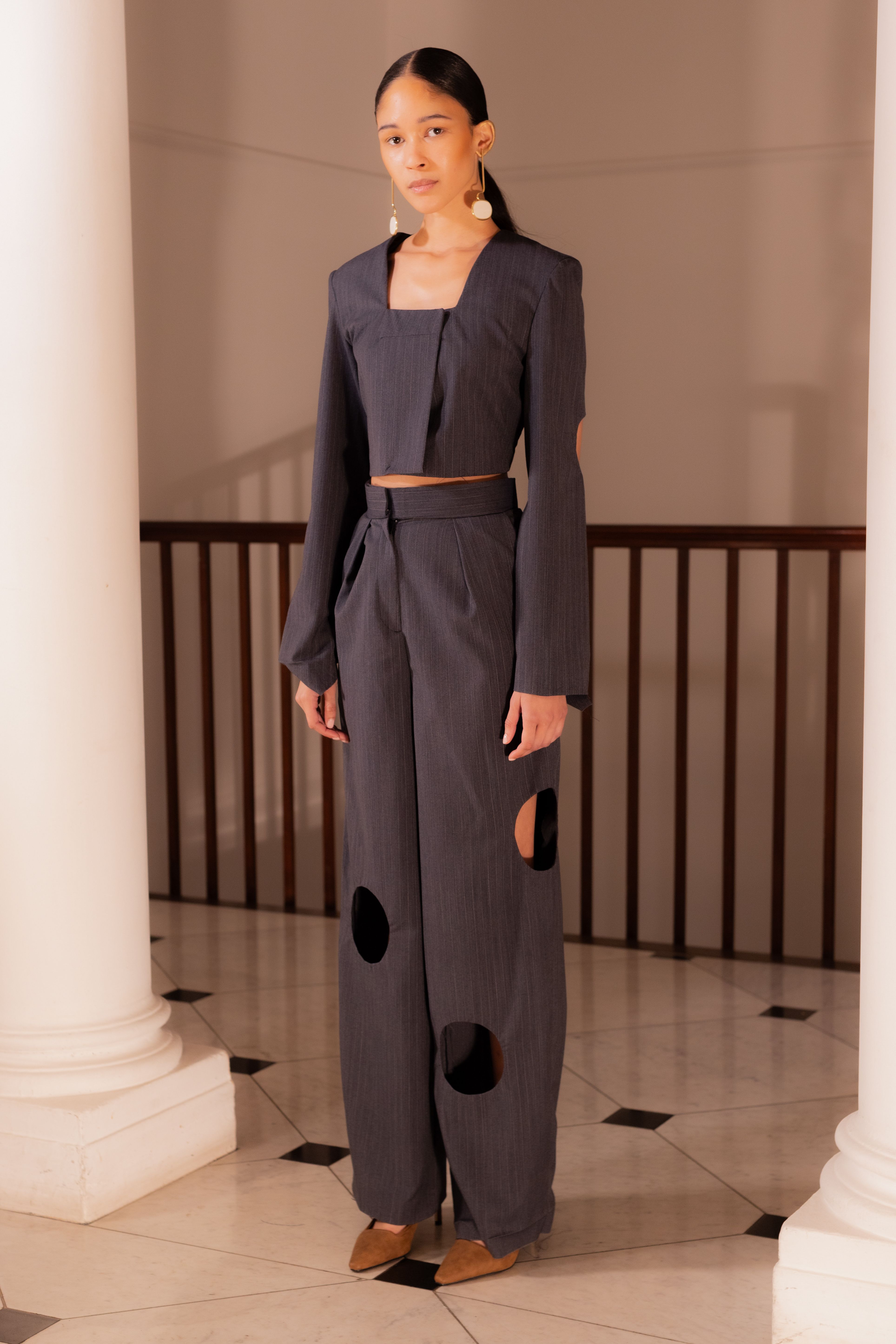 Model wearing a dark grey suit with a cropped jacket and high waisted trousers. The trousers have circle cut outs on the lower leg. 