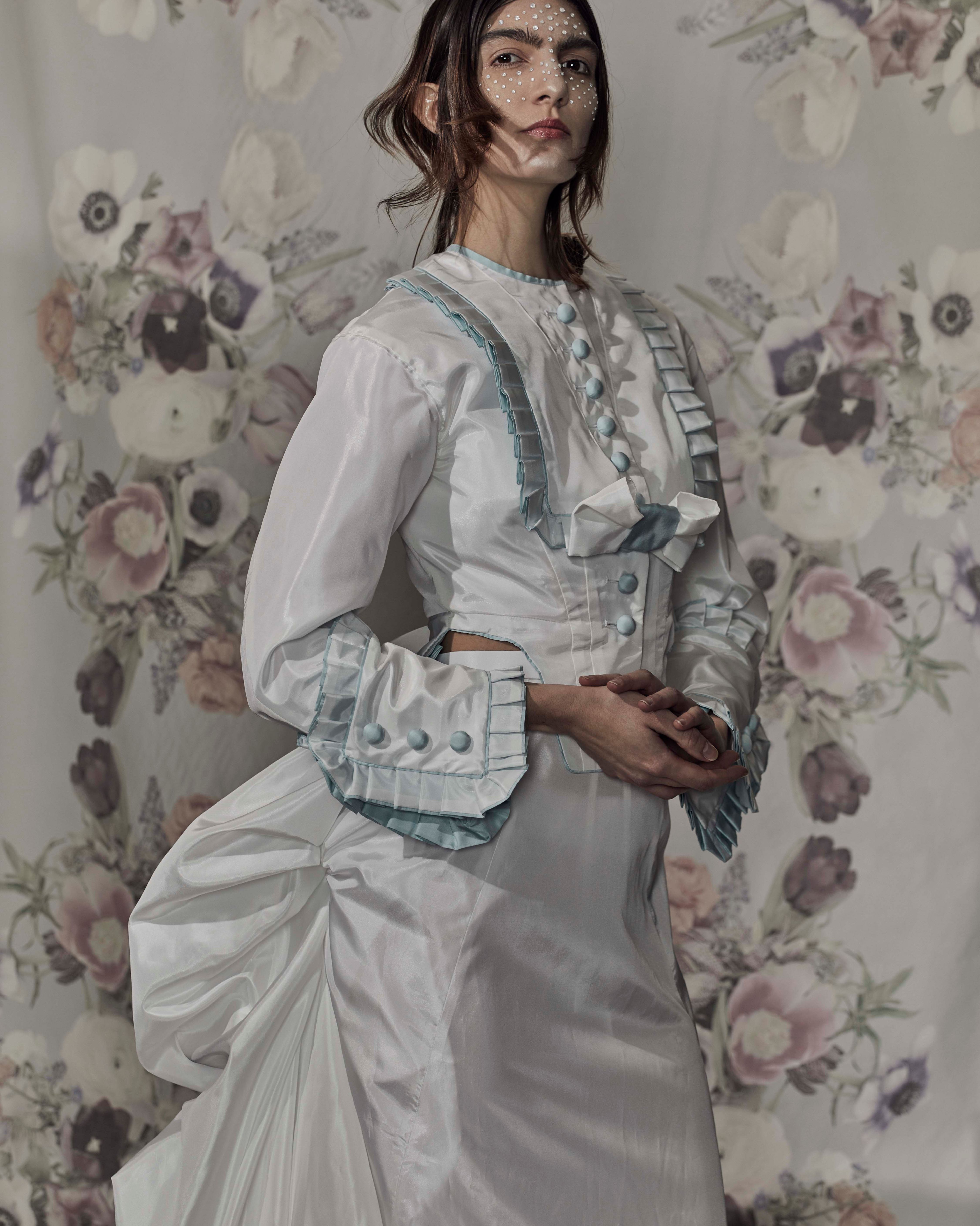 A model in a white, Victorian-inspired dress poses confidently, while a person behind holds a floral tapestry backdrop, creating an artistic and elegant scene.