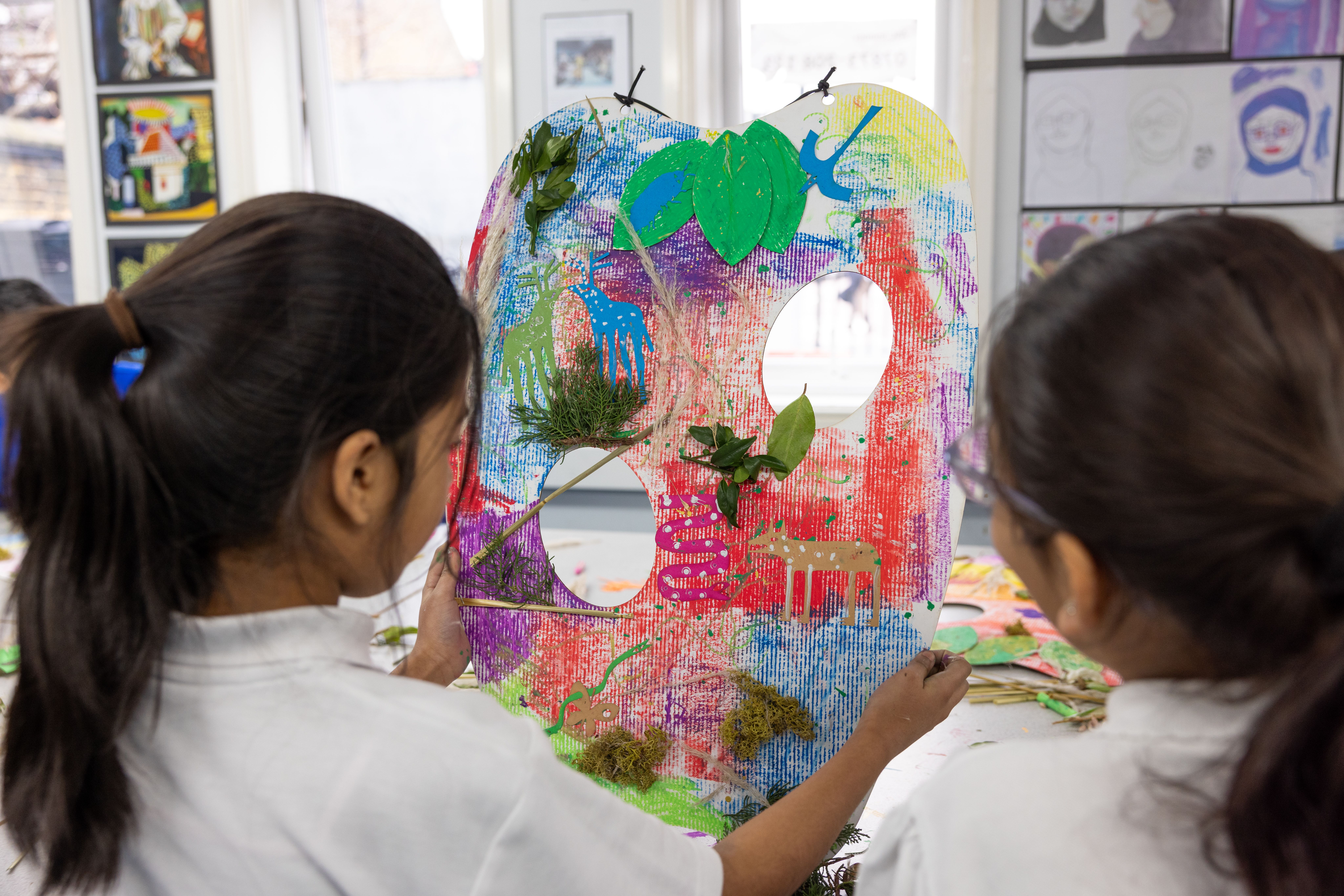 Two school children with ponytails holding a cardboard apple shaped artwork with holes and leave stuck to it.