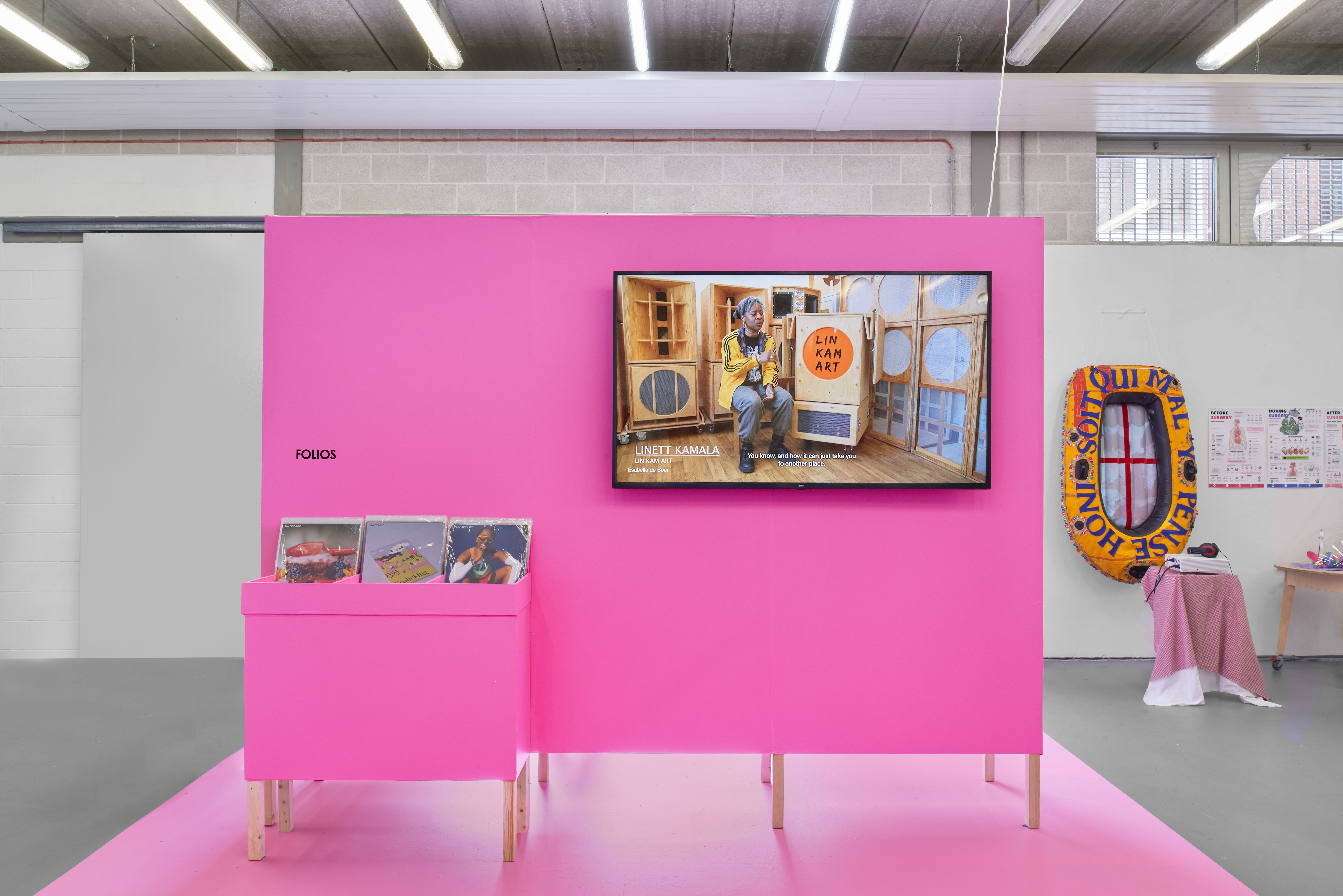 Bright pink exhibition display featuring a wall-mounted video screen and a stand of printed folios in a gallery space, with additional artworks visible in the background.