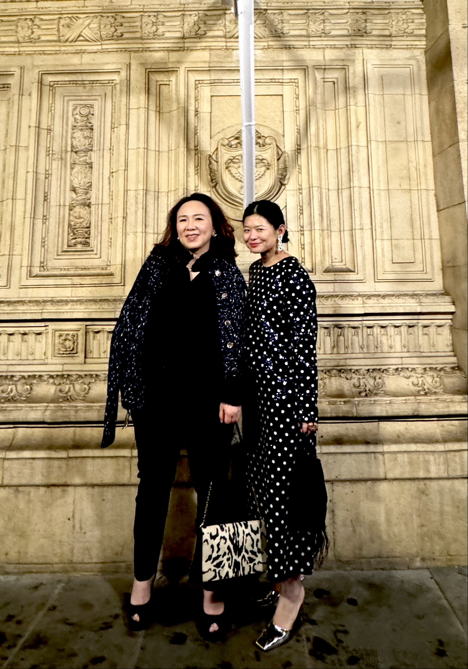 Jasmine and Kuo-Fang stood in front of a stone wall. They're both wearing monochrome, evening wear. 