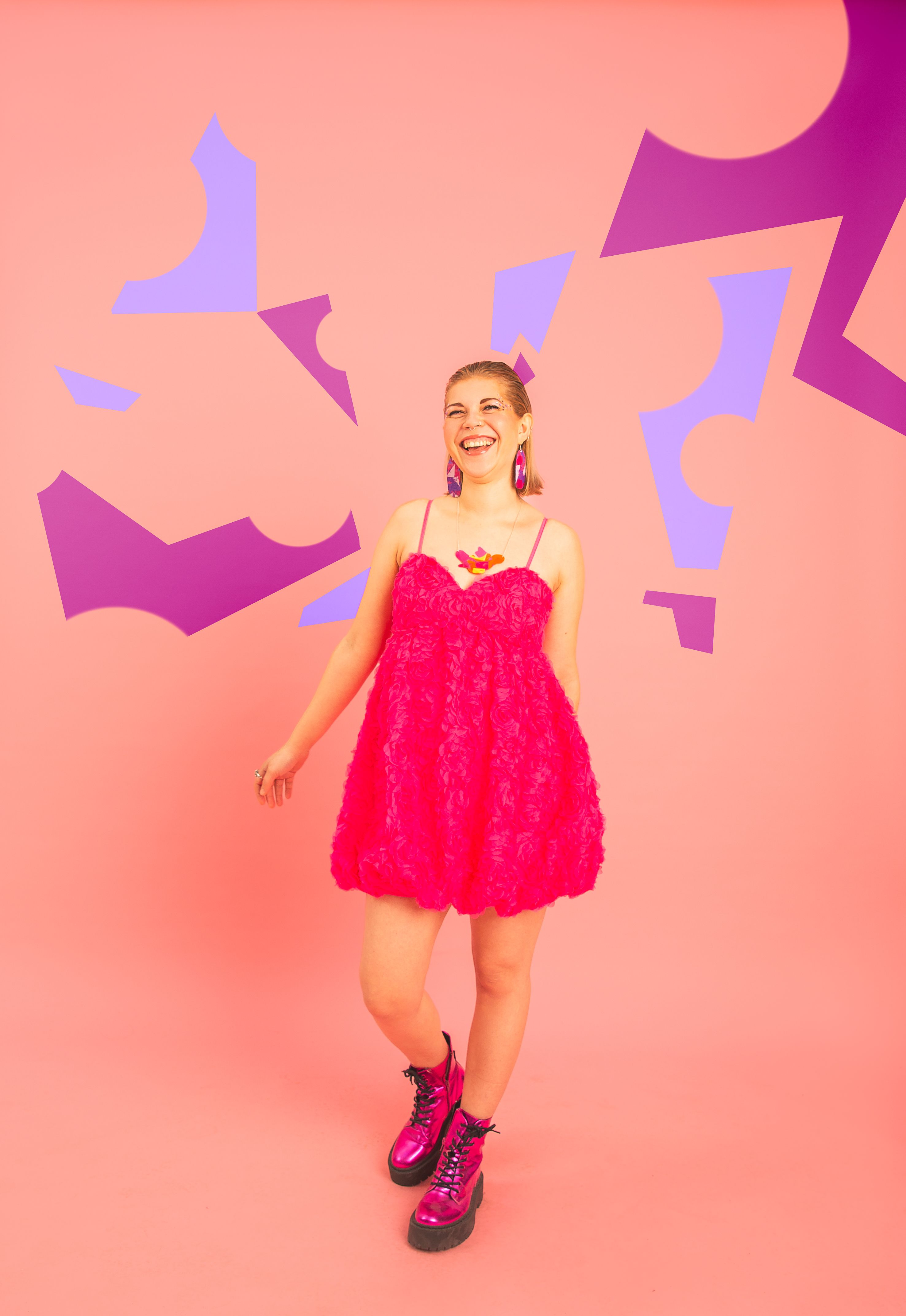 Sian is wearing a pinky mini dress and metallic pink boots. The background is pale pink and there's light and dark purple abstract shapes around her