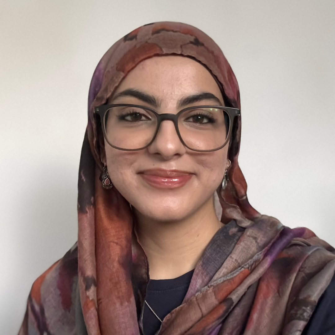 A student smiles at the camera in a head-and-shoulders portrait, wearing glasses and a patterned headscarf against a plain background.