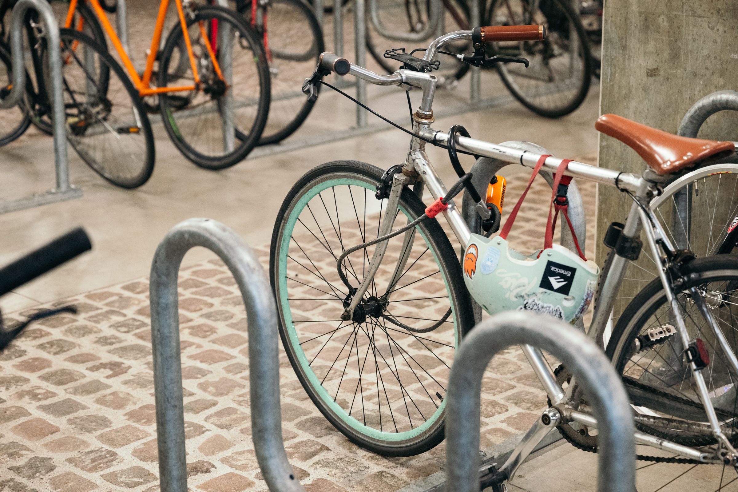 A silver bicycle with a brown seat is locked to a rack in an indoor area. A mint-green helmet hangs on the bike. Other bikes are visible in the background.
