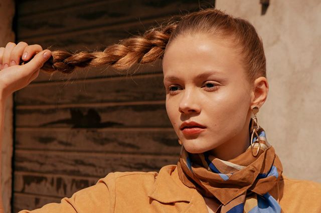 model posed holding the end of their ponytail braided hair, wearing a brown beige and blue scarf and sandy garment