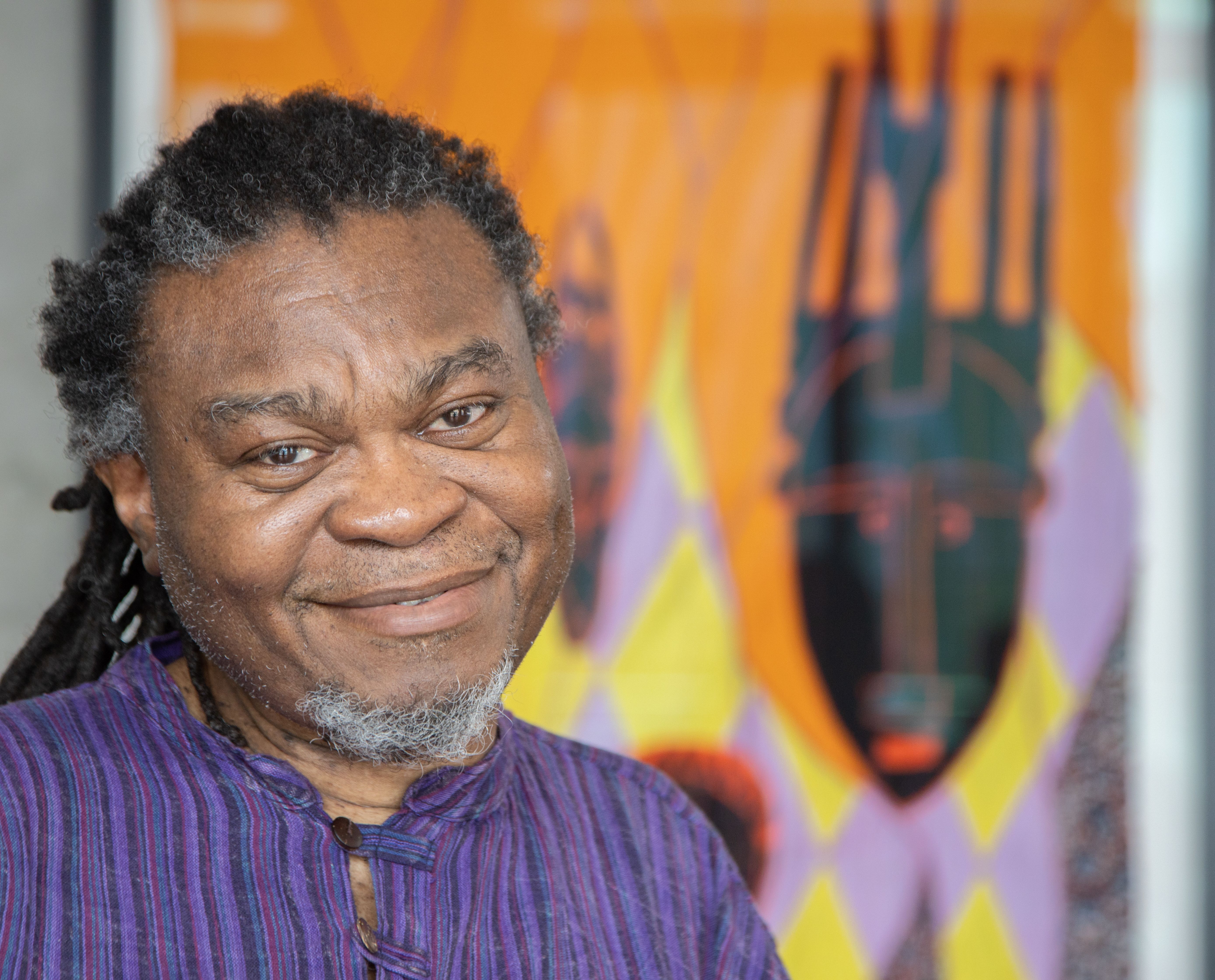 Portrait of Yinka Shonibare CBE RA smiling at the camera. He is wearing a purple shirt and the background is out of focus.