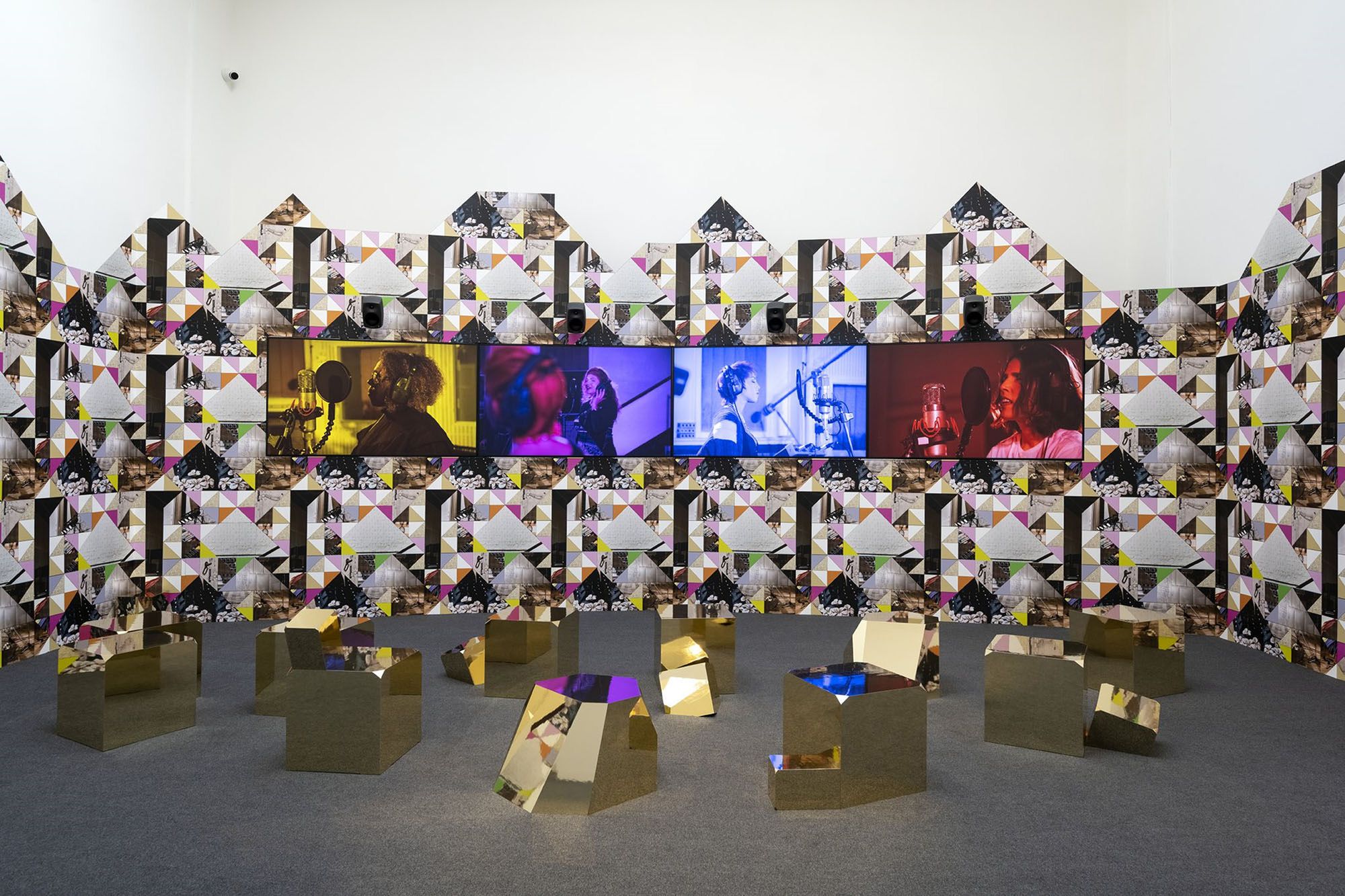 Image shows a digital render of Sonia Boyce's Venice Biennale exhibition, Feeling Her Way. The image shows angular gold blocks sitting on the ground. In the background are several screens with images of people singing and performing music. The screens sit on a multi-coloured triangular wall. 