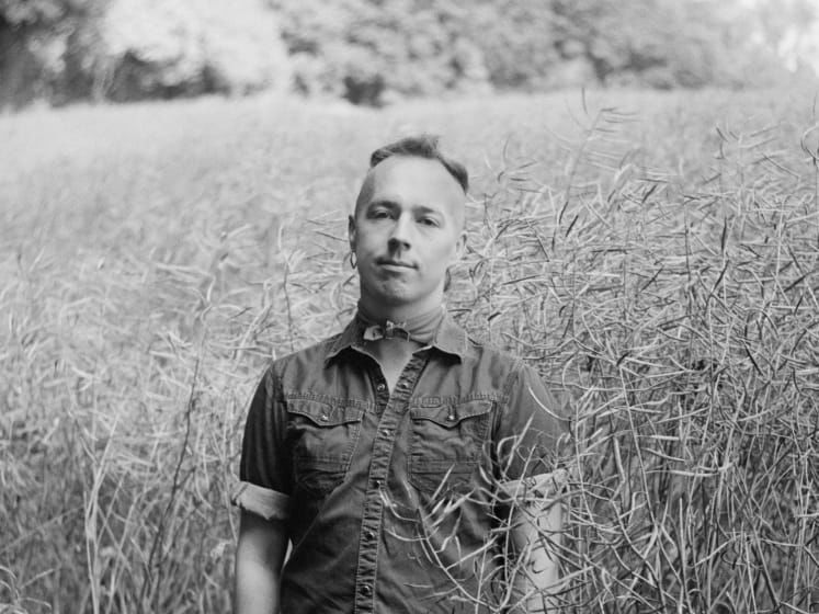 A black and white photograph of a person standing in the middle of a field with plants surrounding them. They are wearing a short-sleeved denim shirt a neck tie and earrings. 