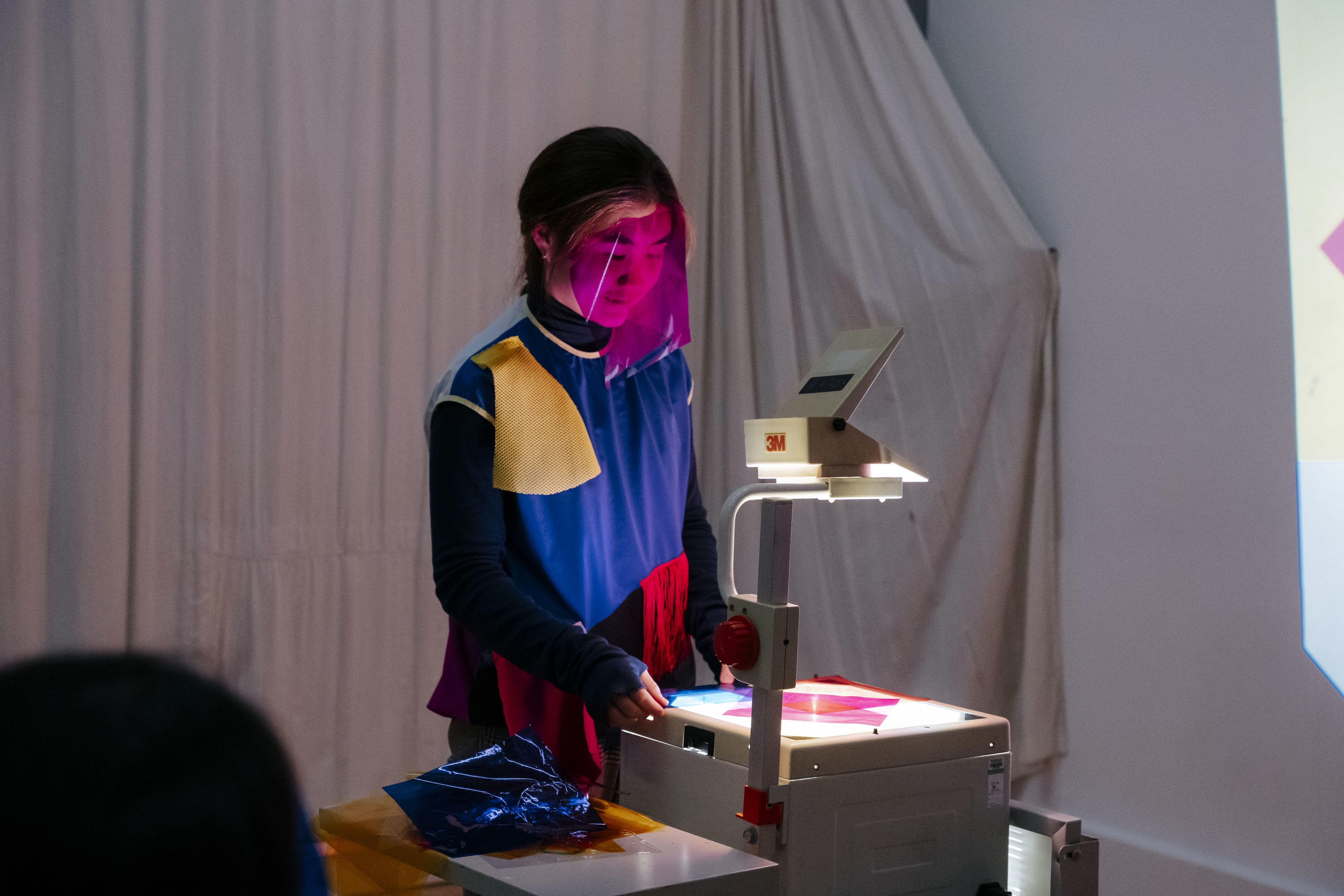 A person is standing in front of an overhead projector, manipulating colored transparent sheets. They are wearing a multicolored outfit and have a pink transparent sheet covering their face. The setup suggests they are creating a visual display or presentation, possibly for an art performance or educational purpose. The background features white drapes, contributing to a focused and minimalist setting.