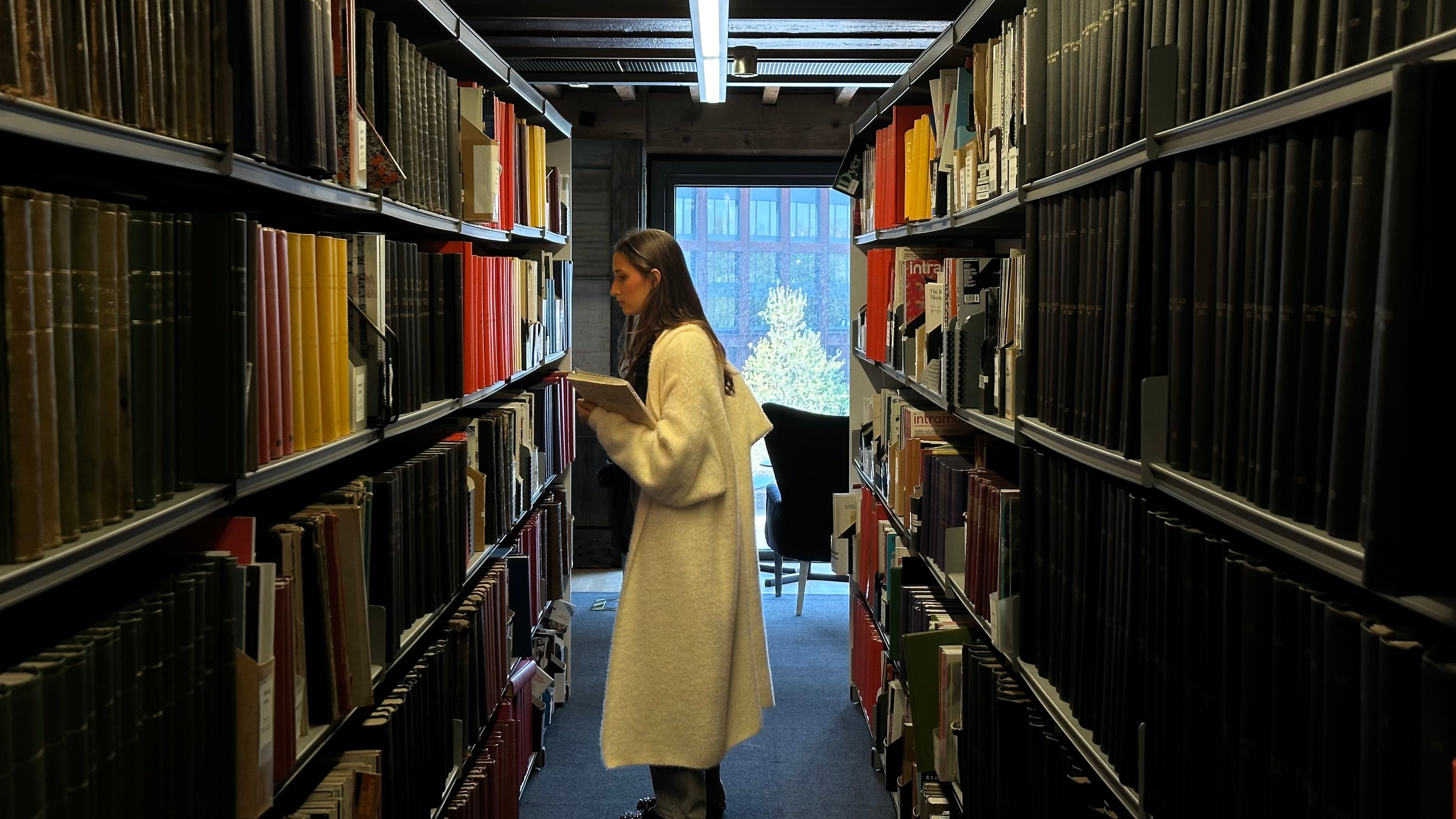 Violetta, holding a book, looks through the library shelves 