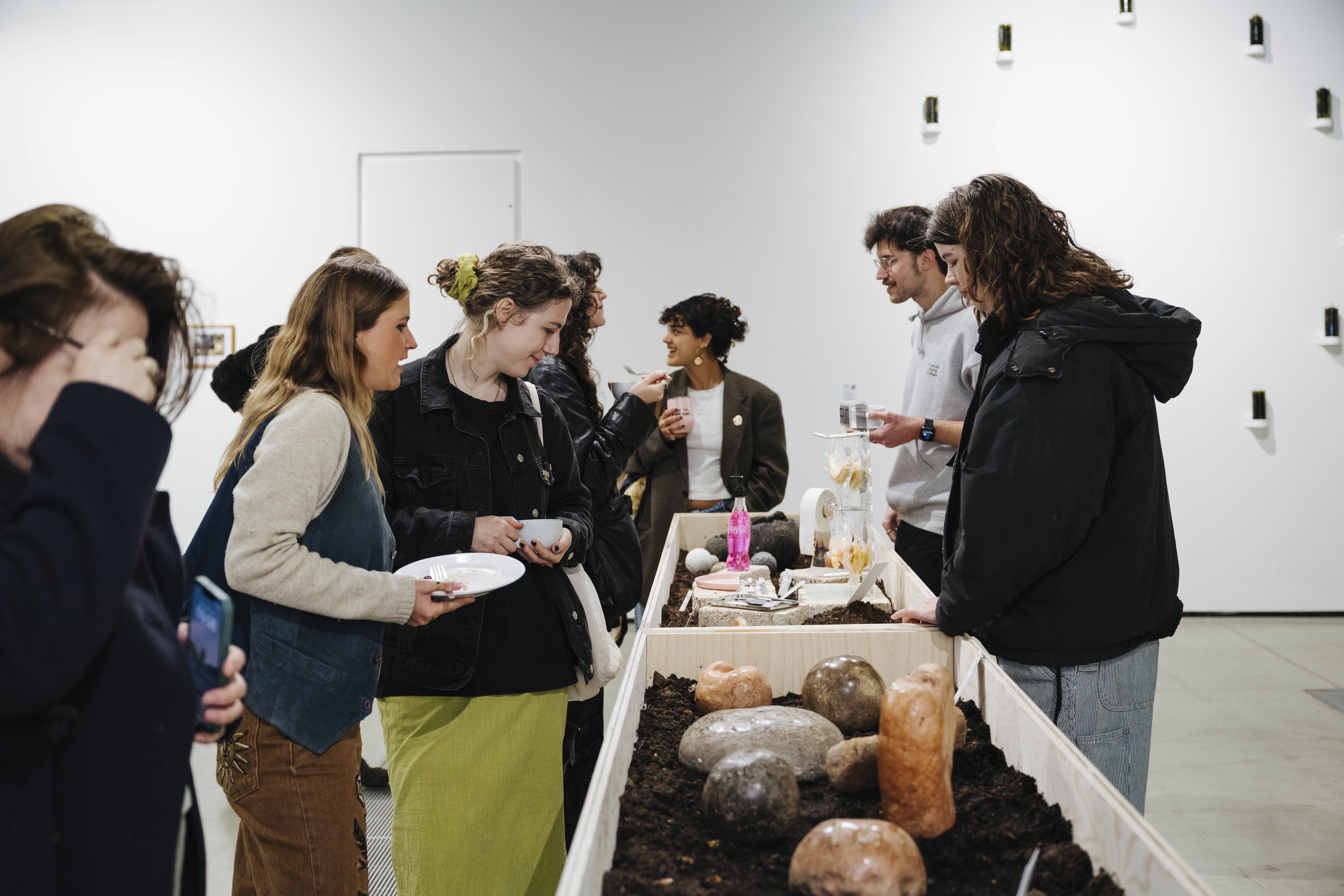 A group of people standing around a wooden open-topped vitrine in an art gallery. They are looking into the vitrine at exhibits which are placed in soil.