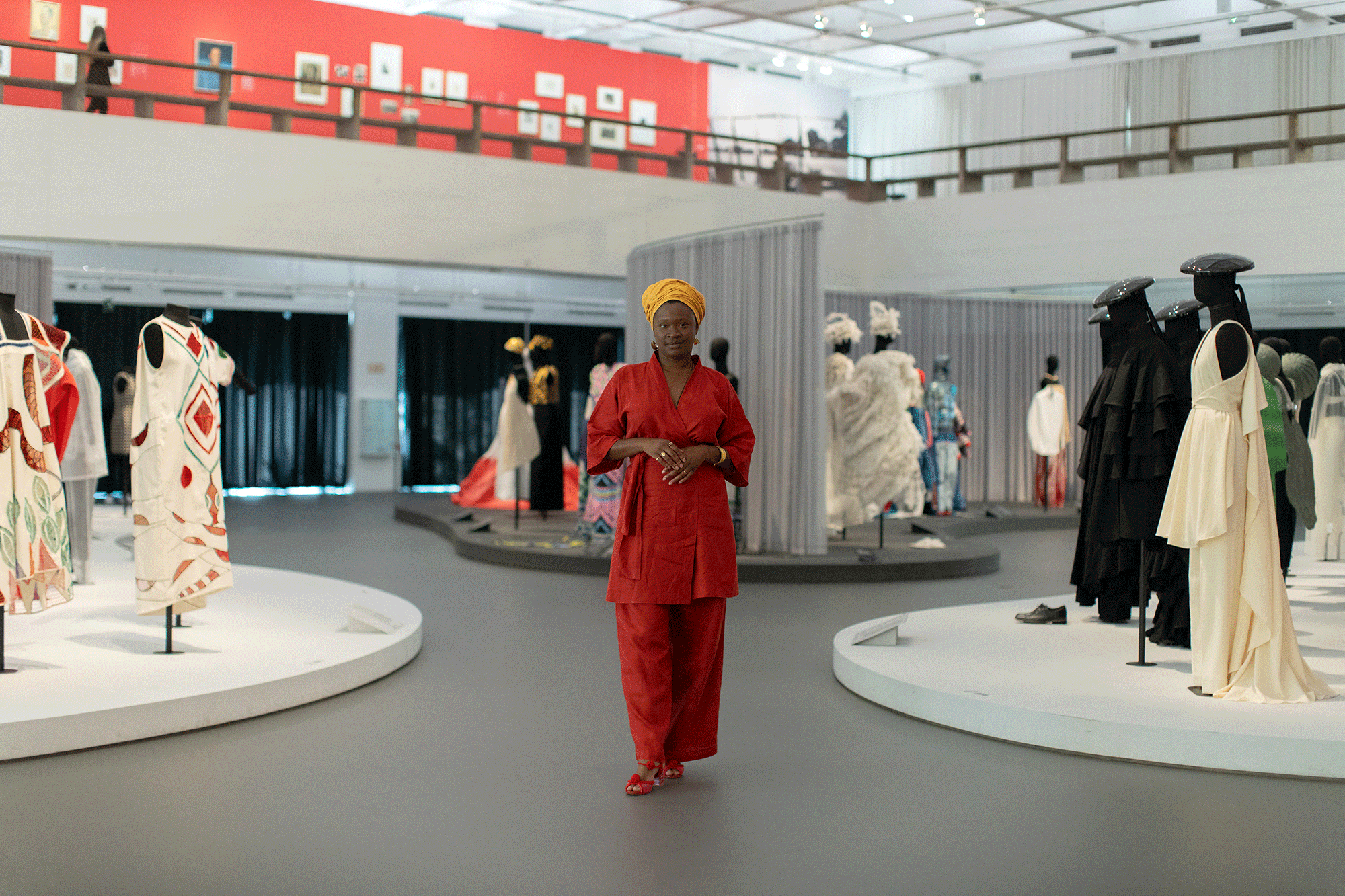 Image shows a Black woman standing in an exhibition space with fashion bodices scattered in the background. She is wearing a red outfit with a yellow headpiece.