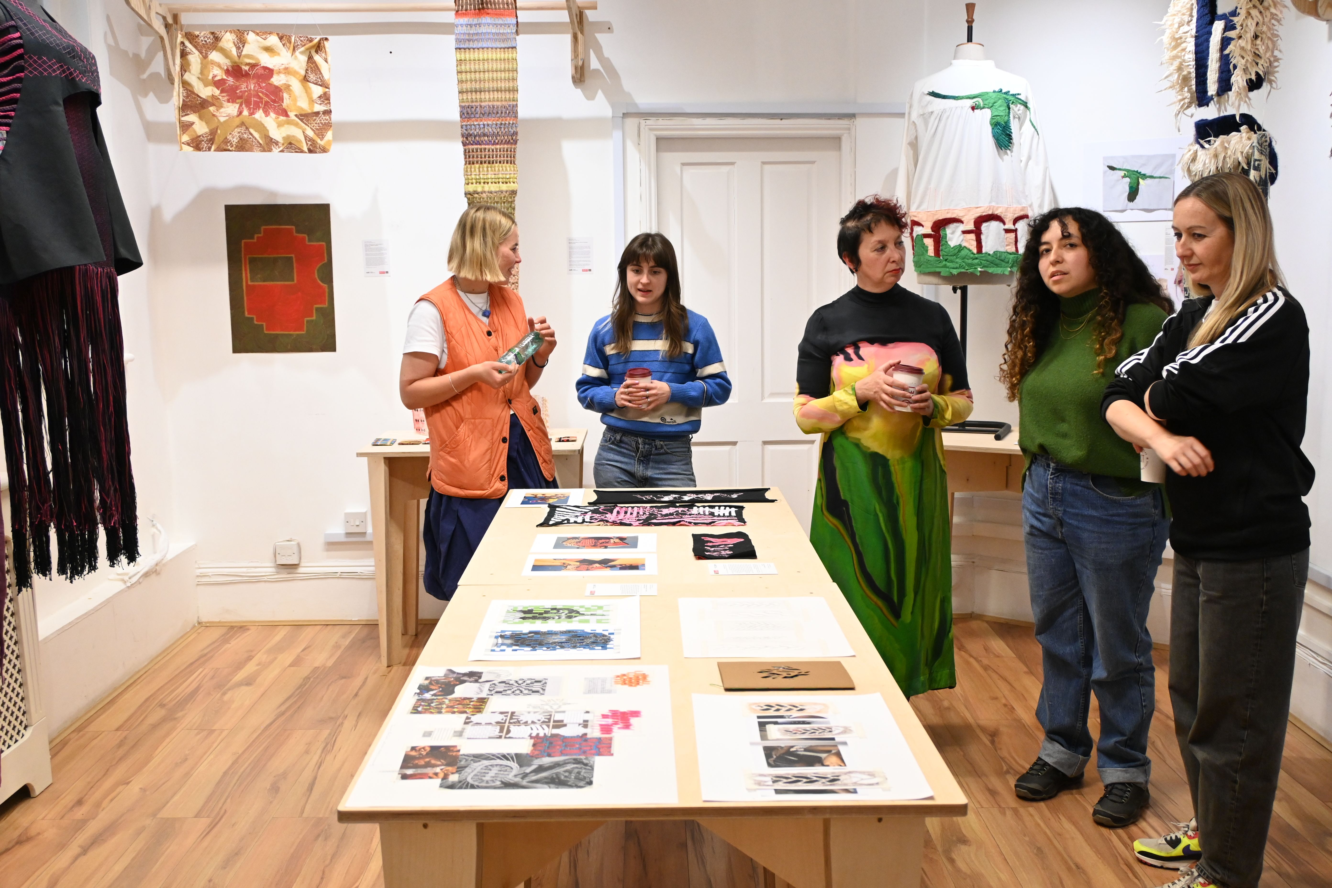 Image shows 5 people standing round a table with textile-based artwork in the background and on the table. The people are engaged in discussion as they admire the work. 