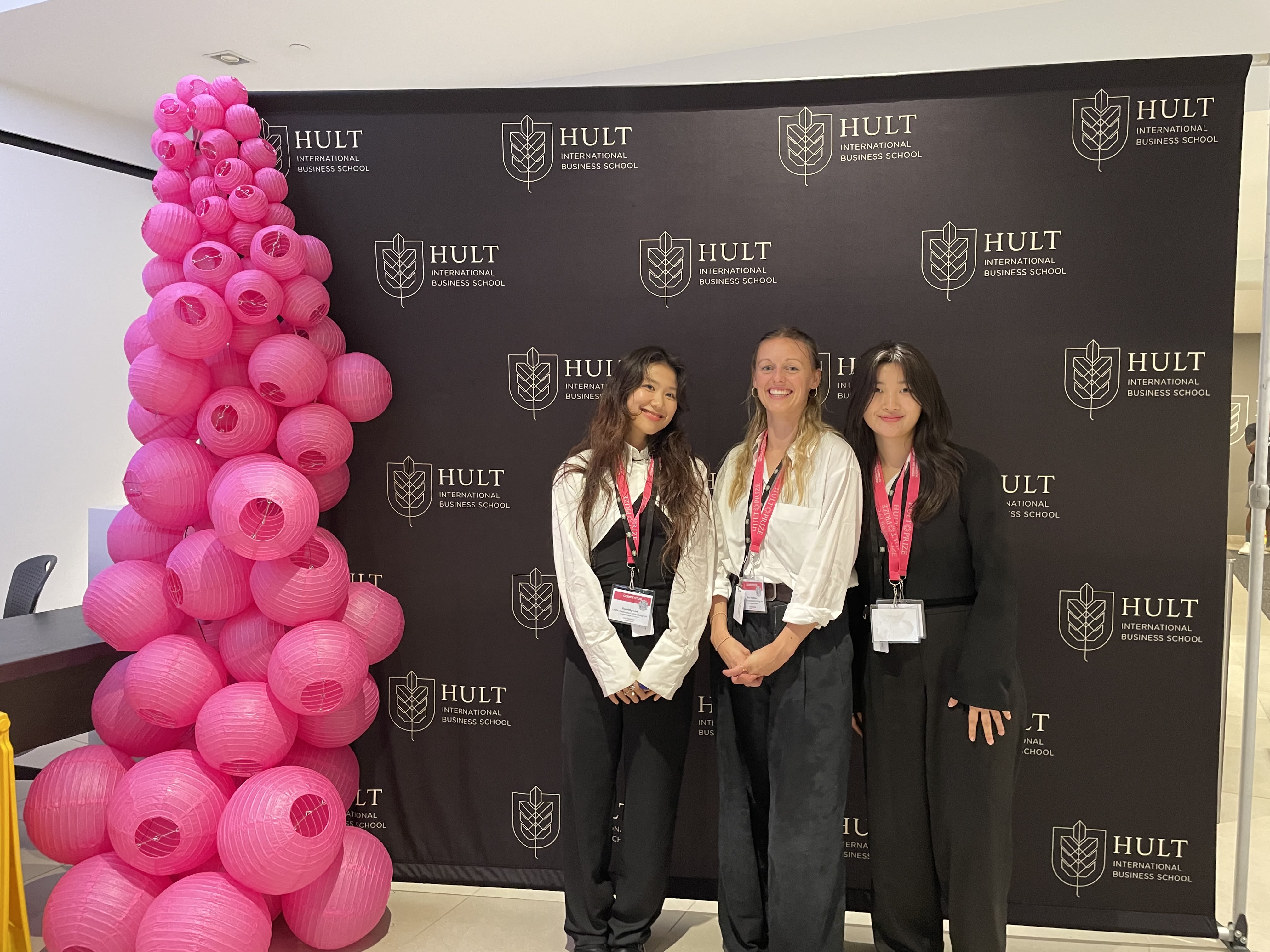 Three people standing against backdrop with 'Hult' Branding, next to a stack of pink balloons, smiling at camera.