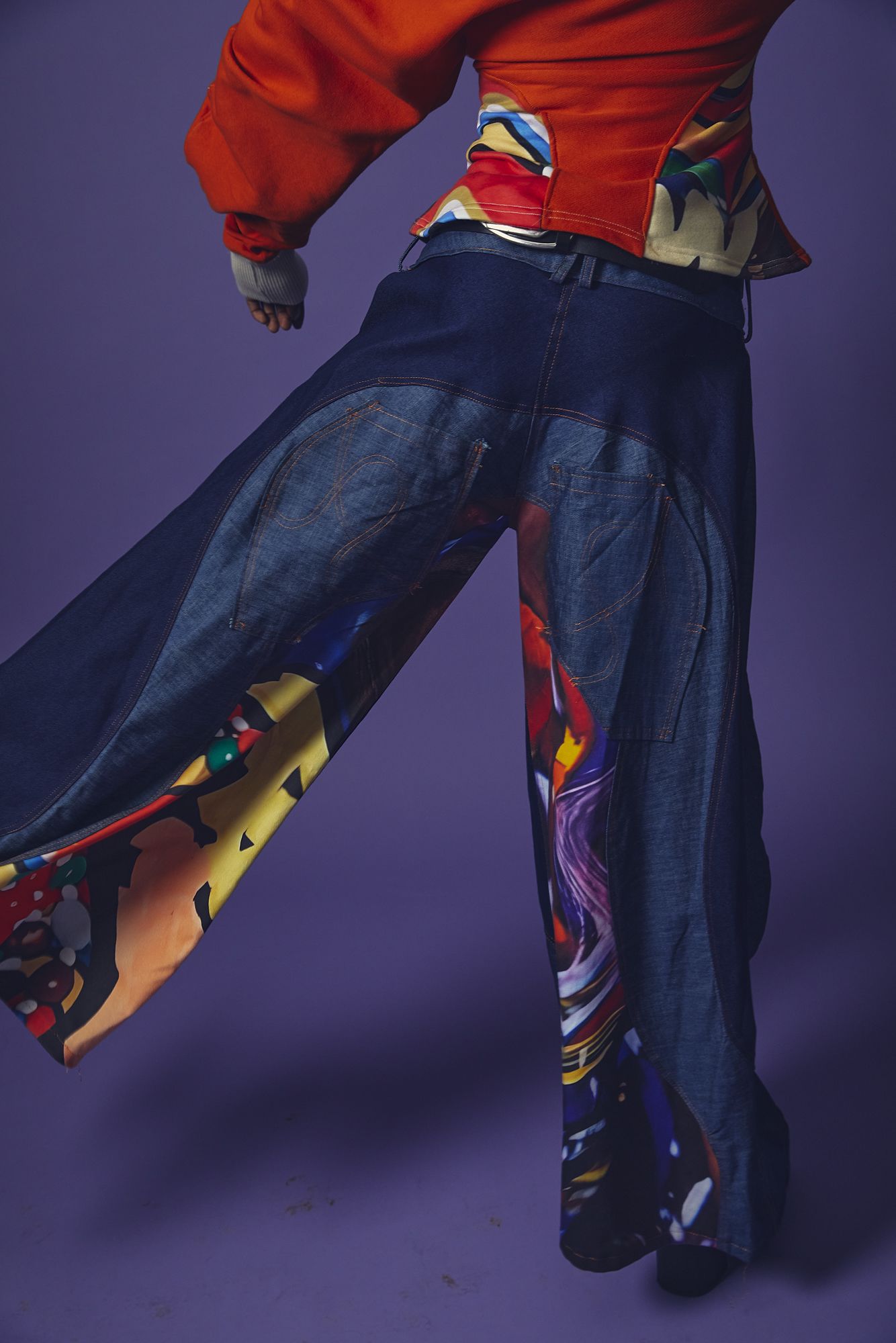 Person wearing wide-leg denim jeans with colorful printed fabric inserts and an orange top, posing against a purple background.