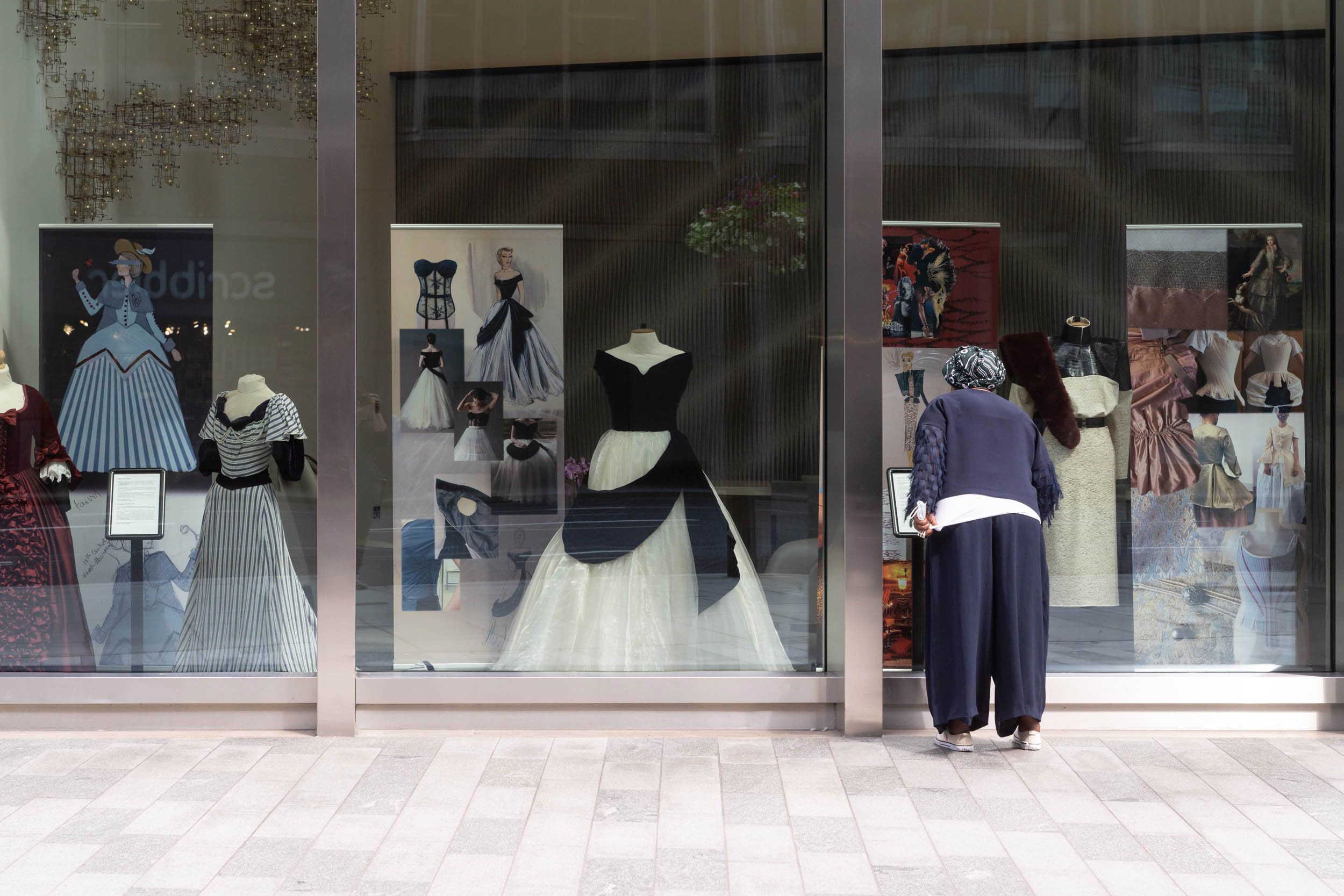 A woman with her back to the camera peers into a large shop window where three elaborate costumes are on display