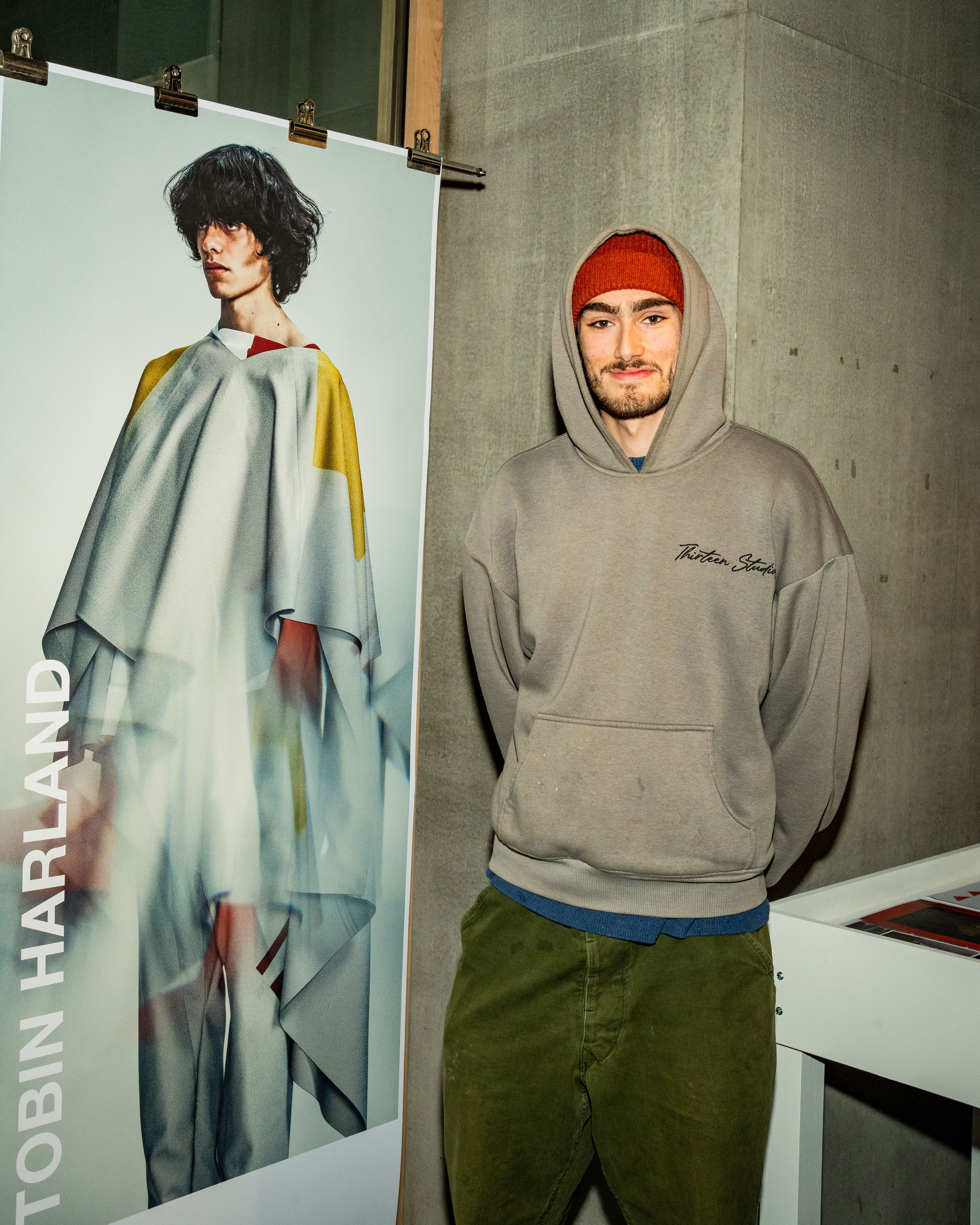 Person smiling at camera, next to roll down poster of model dressed in grey garment.