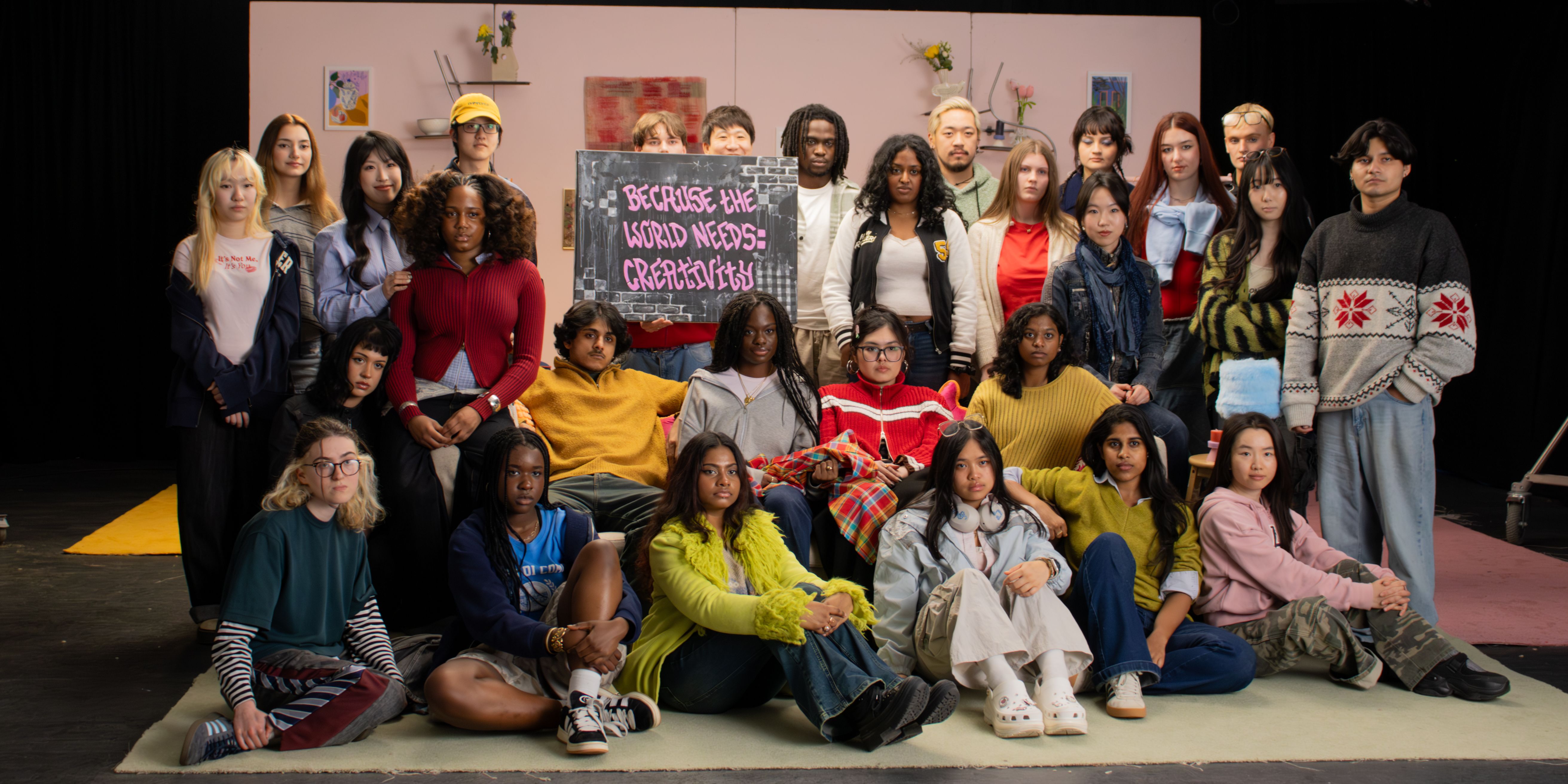 A large, diverse group of approximately 25 young people — students of various ethnicities — are arranged in three rows on a studio stage set. The backdrop is a dusty pink flat decorated with small framed artworks, shelves with ceramics, and dried flower arrangements, suggesting a lived-in domestic space. In the centre of the group, one person holds up a hand-painted chalkboard sign in pink graffiti-style lettering that reads: 