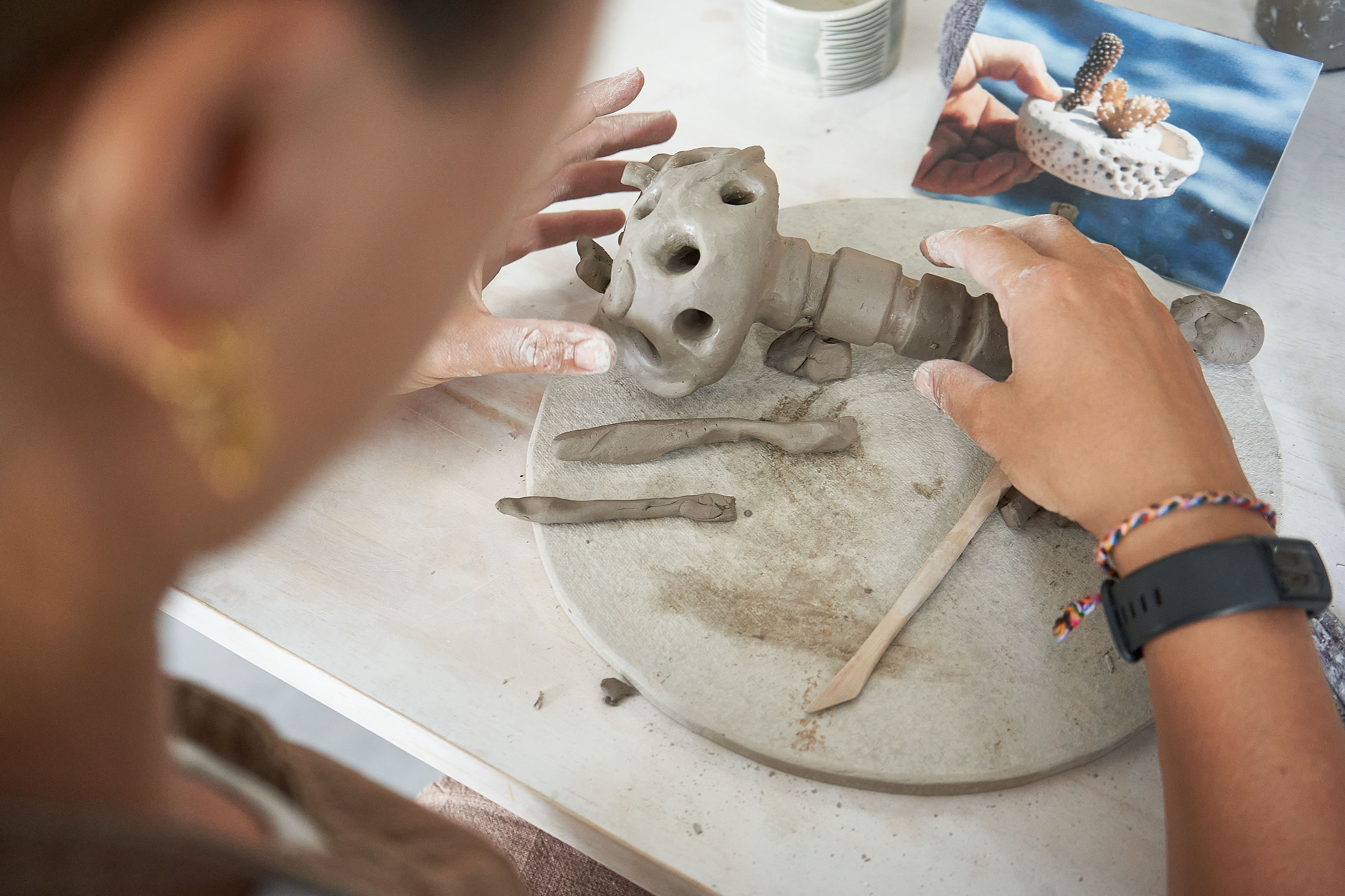 Close-up of two hands shaping a small, textured clay form with holes on a circular work surface, with clay tools and rolled pieces nearby; a printed reference photo of a coral-like ceramic object sits in the background.