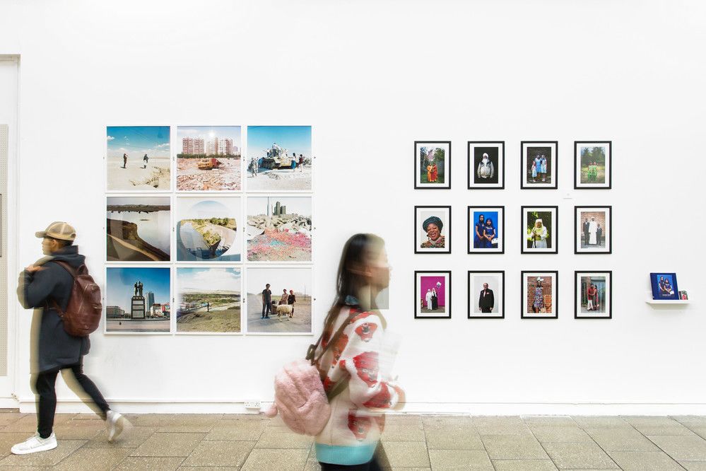 Two people walking in opposite directions past a white wall with framed photography as part of an exhibition. The shot is slightly blurred.