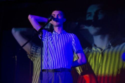Photograph of a man in a striped shirtspeaking into a microphone on stage, one arm raised behind his head
