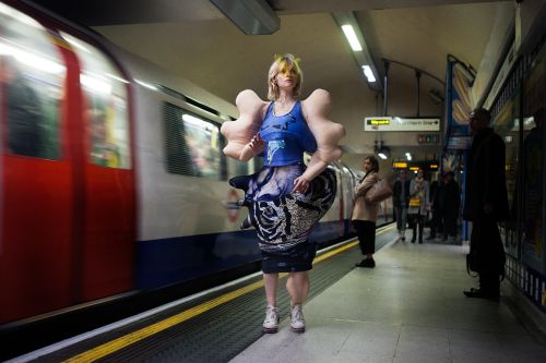 model at undergound station beside a moving tube, wearing a bodysuit garment with disfigured limbs