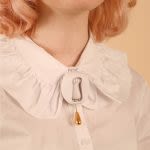 Photograph of a close up of someones chest, showing a collar brooch against a frilly white shirt collar, the brooch is circular and shows a cat's paw with a little bell dangling below
