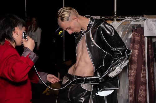 Male model with a shaved head wearing a black open PVC coat having his belt put on by a designer