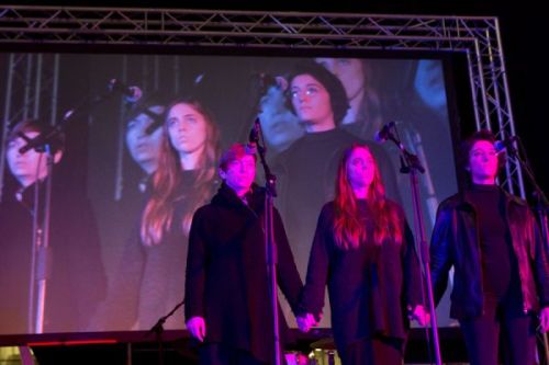 A photograph of three women standing on a stage holding hands under pink/purple lighting, an image of them projected onto a screen behind them