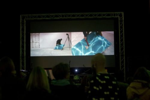 A photograph from behind a crowd watching a large screen above a stage as someone performs with a flat, square blue object on a split screen projection