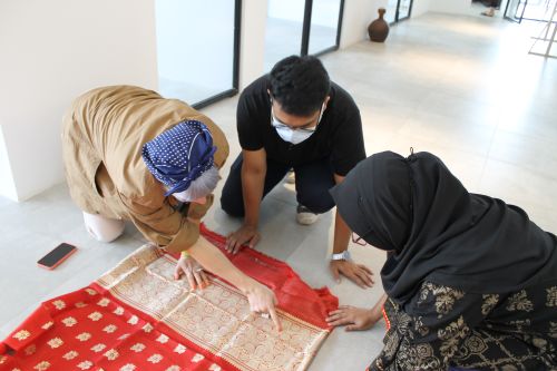 3 people kneeling over a piece of printed fabric. One is wearing a mask and 2 are wearing headscarves.