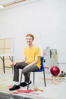 A young man in a yellow t-shirt sat on a chair in a painting studio 