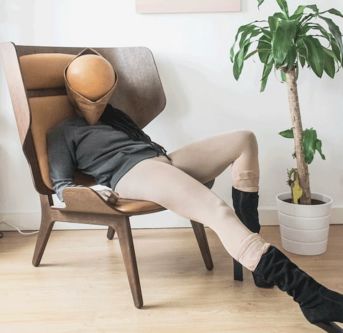A figure reclines on a chair with a pot plant, their face is an orange ball beneath a hood