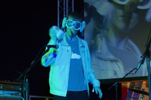 A photograph of a man on a stage under heavy blue lighting, wearing large goggles and shaking something in one hand