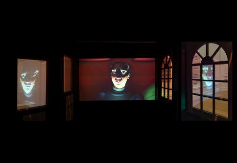 A dark room with a screen in the centre projecting a close-up colour saturated image of a face to the right and left. 