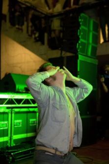 A photograph of someone on their knees, their shirt unbuttoned and they are covering their face with their hands and looking upwards