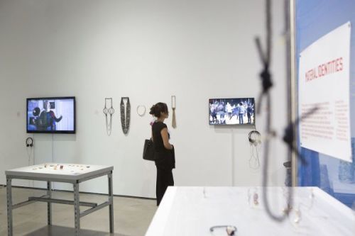 Photograph of someone looking at work installed in the gallery, two televisions hang on the wall in background and a table displays artwork in the middle