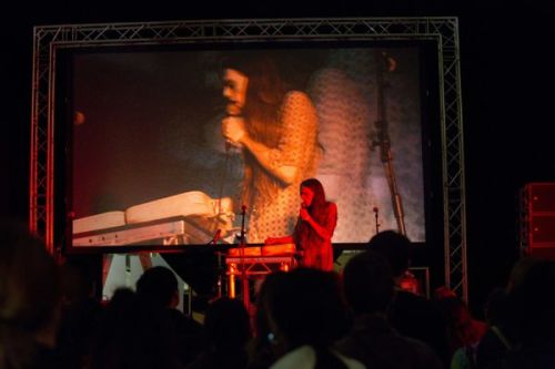 A photograph from behind a crowd watching a woman sing into a microphone on a stage, an image of her is projected onto a large screen above the stage 