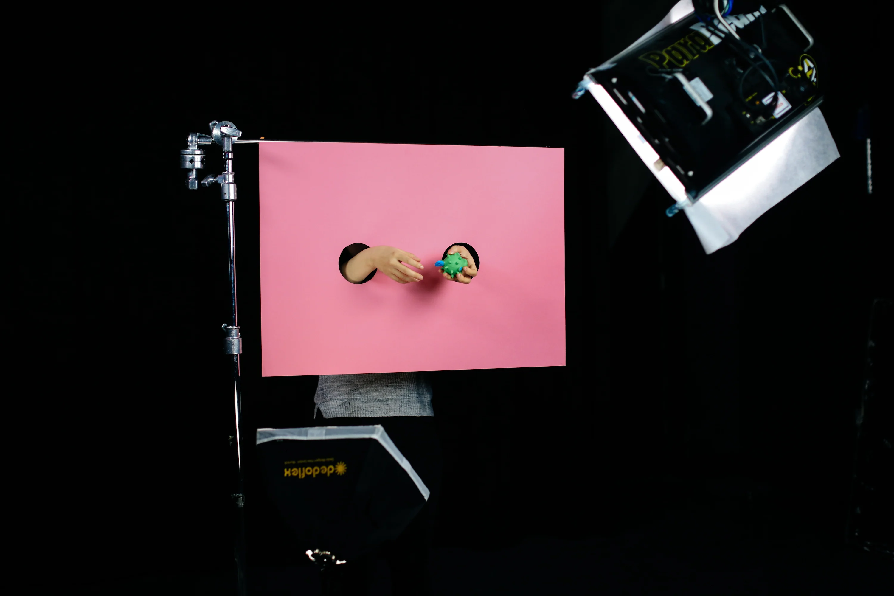 Two hands emerging through hole in a pink screen holding a green item