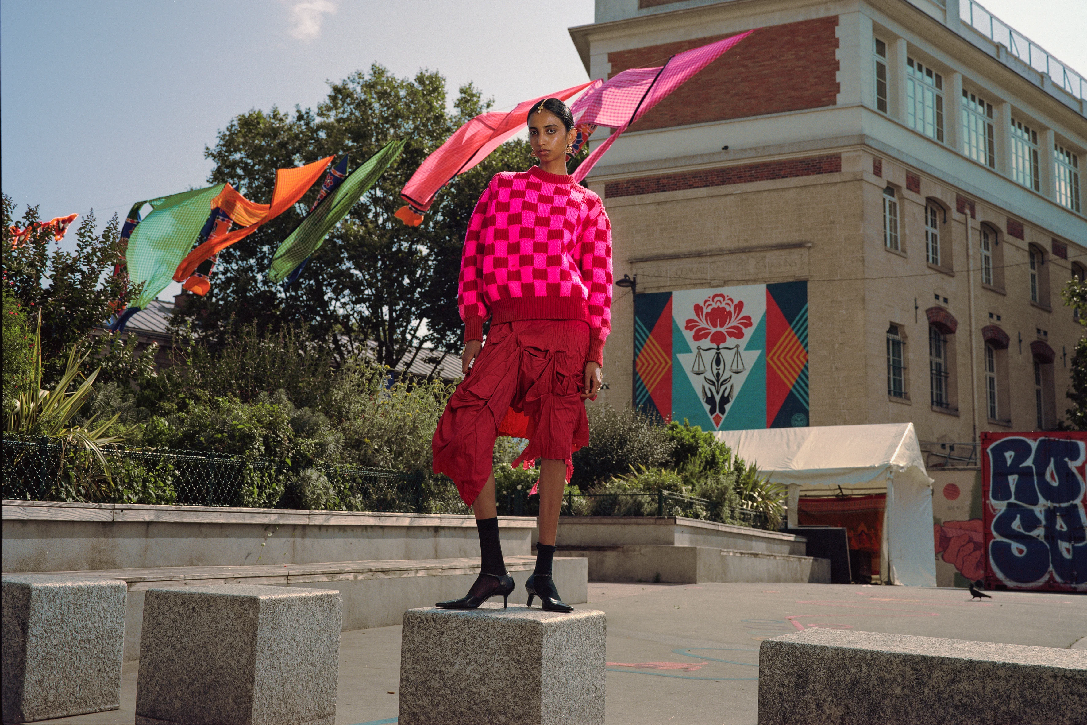 A model in bright pink skirt and patterned jumper is standing on a square marble platform next to a large commercial building just visible. There is greenery in the background and colourful gingham fabrics hanging on a line overhead.