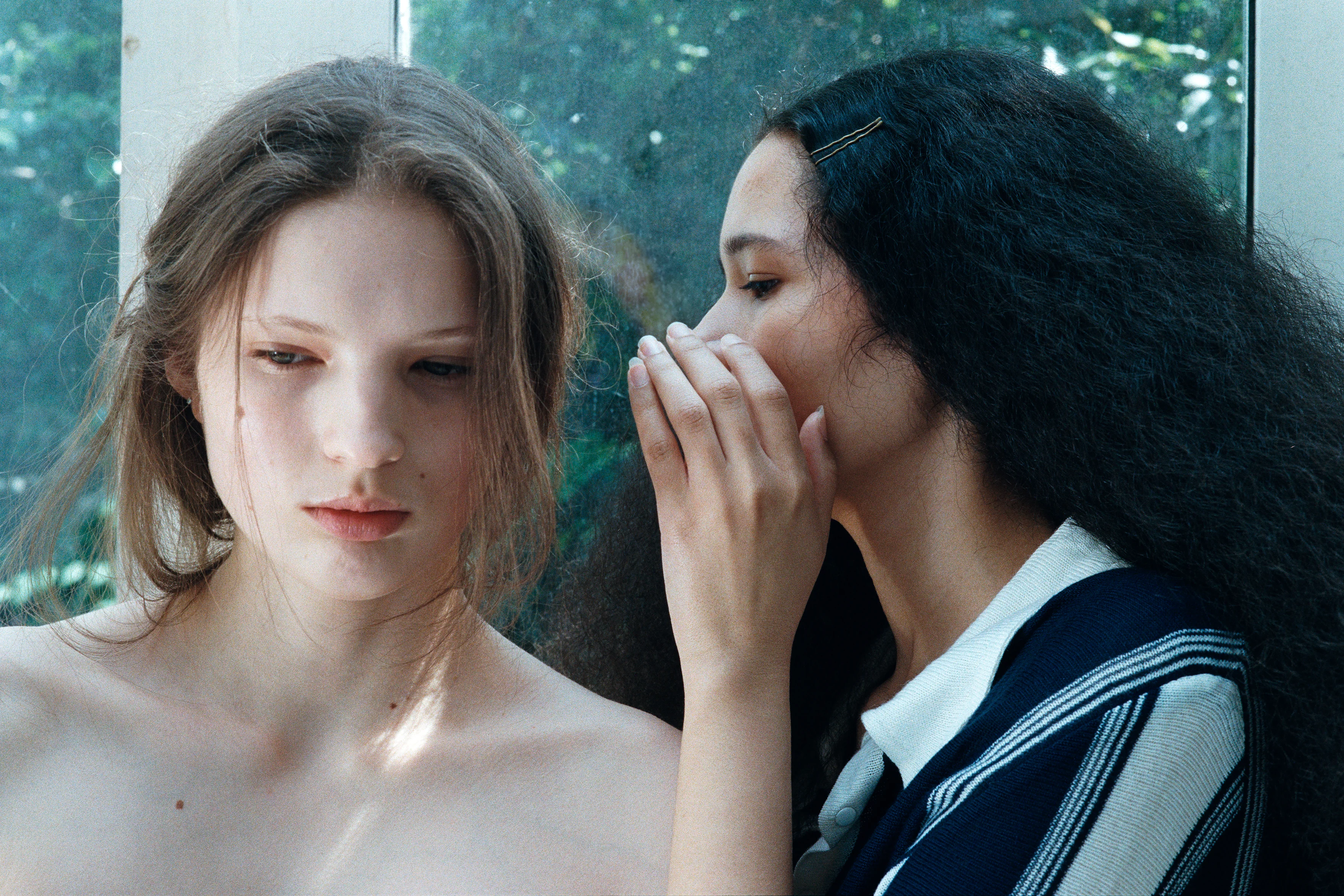 A fashion portrait with two models, one facing the camera, the other has her head turned towards her and is whispering behind her hand. We can see greenery via a pane in the background.