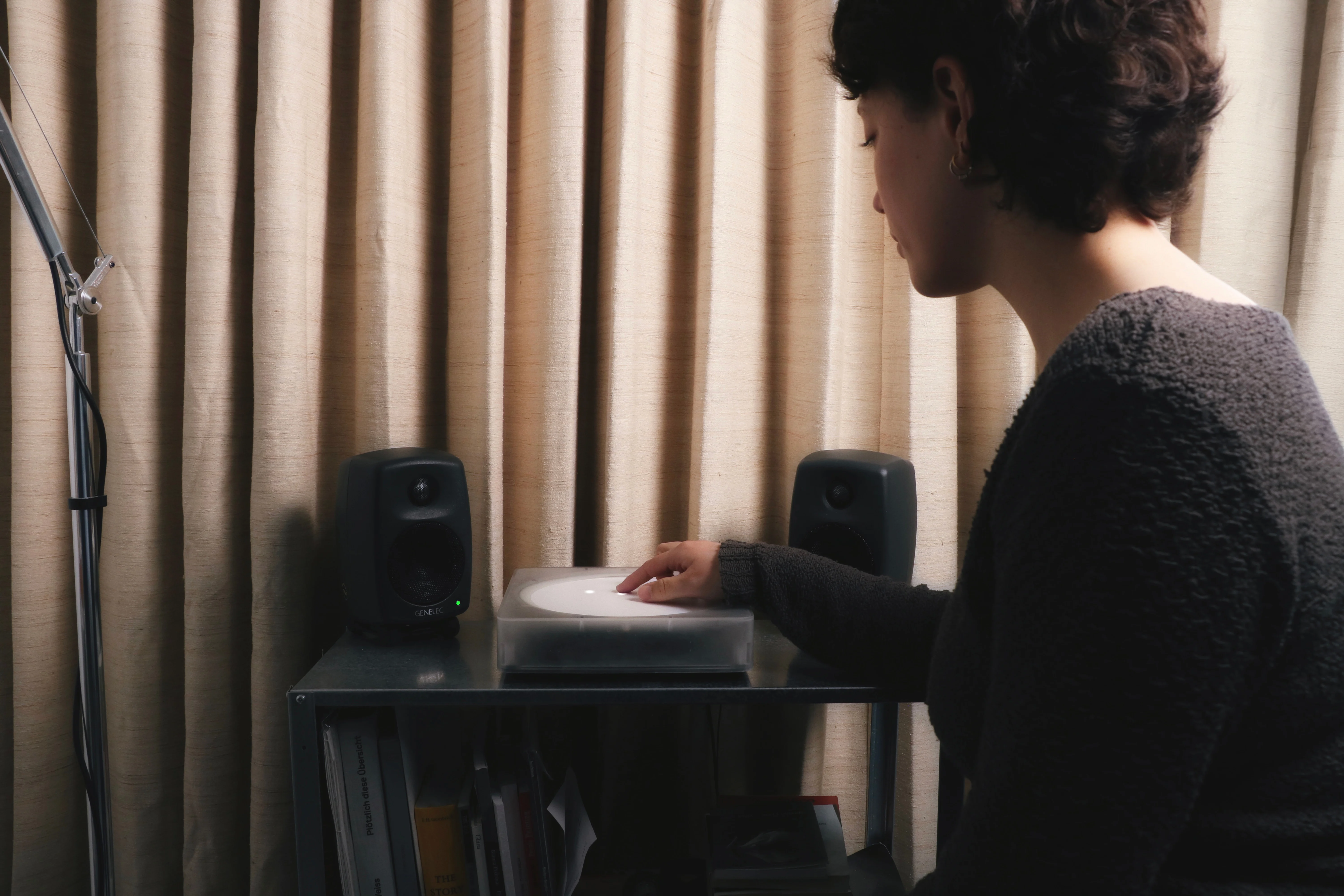 A person is interacting with a small digital turntable on a stand, with speakers on either side. There is a soft draped curtain behind and a lamp stand just visible to the left.