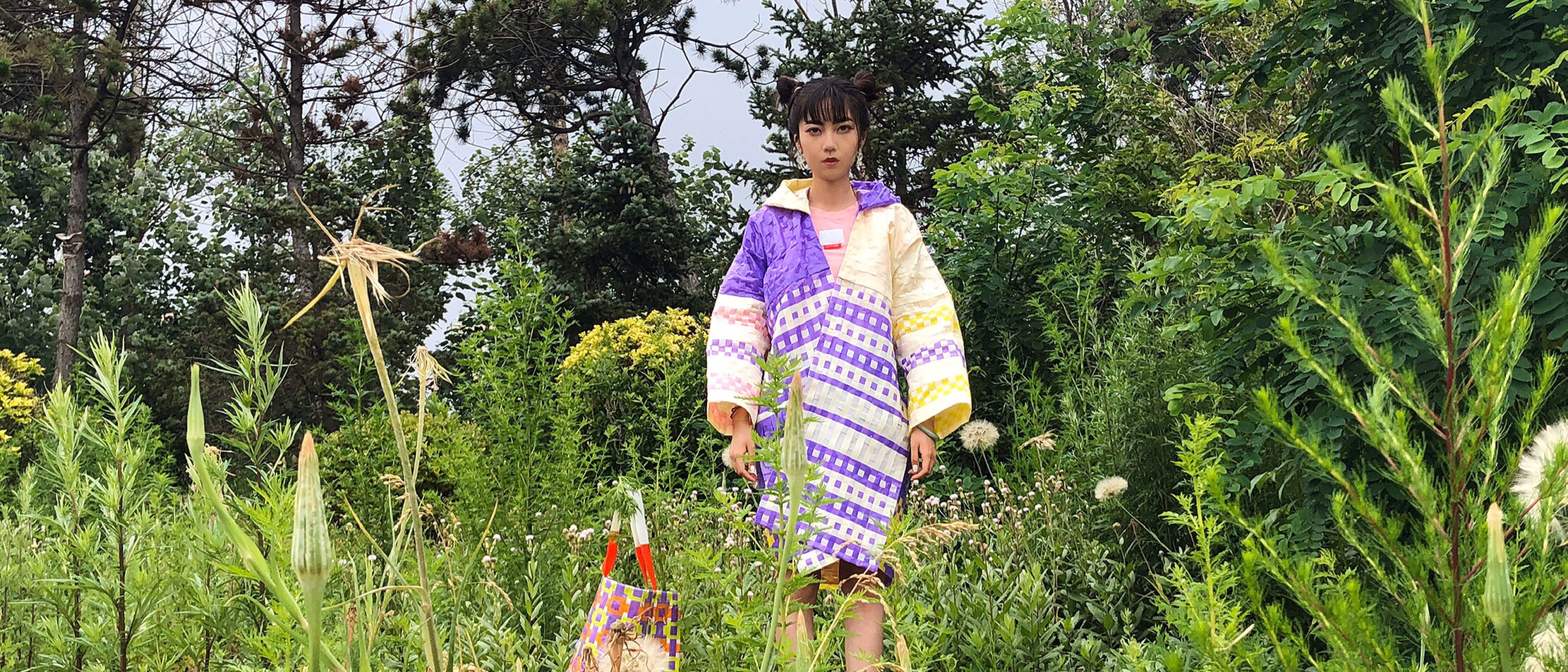 Girl standing amongst grass, tress and bushes wearing a brightly coloured coat.