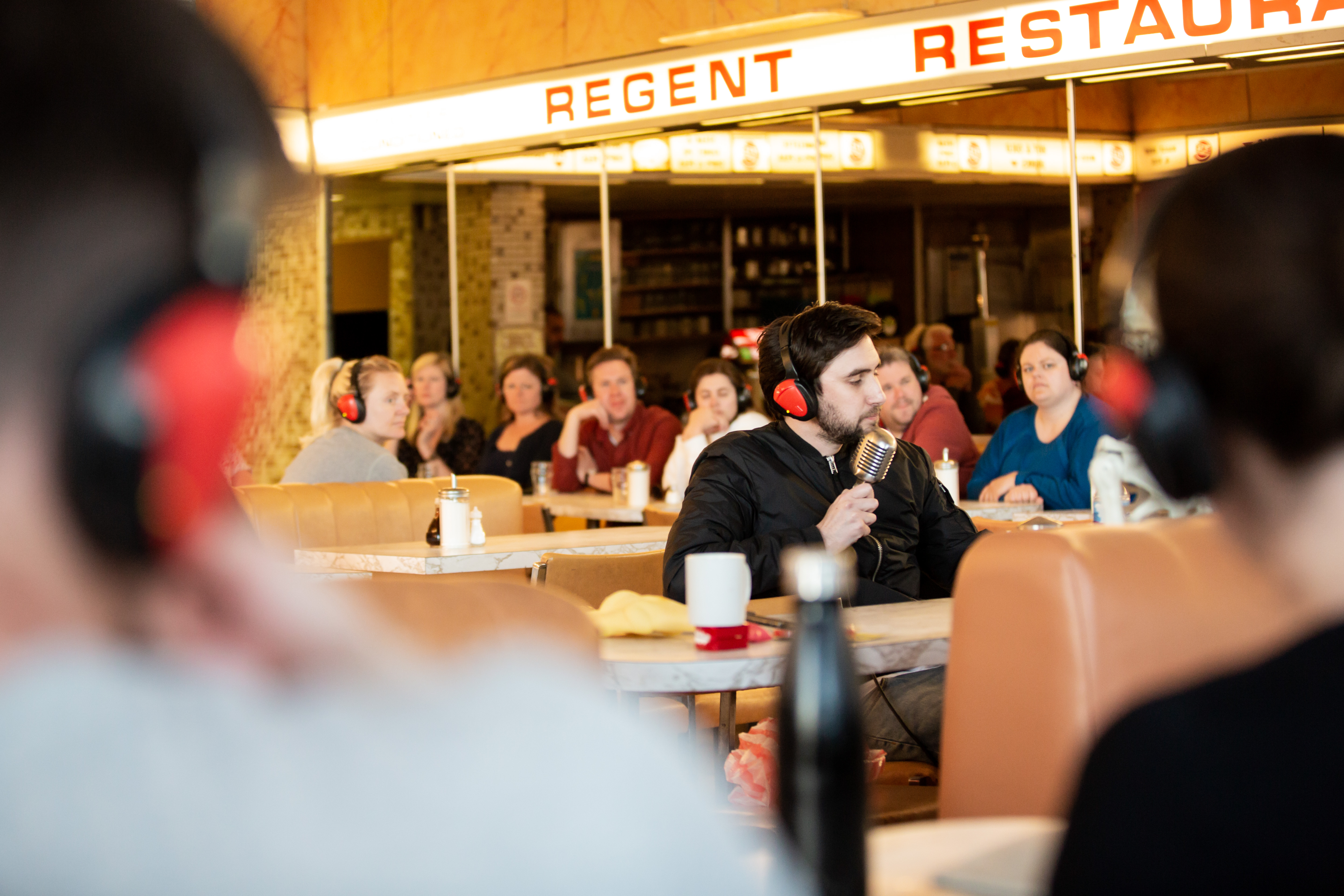 A man holds an old-fashioned microphone in his hand, sitting at a table in a full diner style restaurant.
