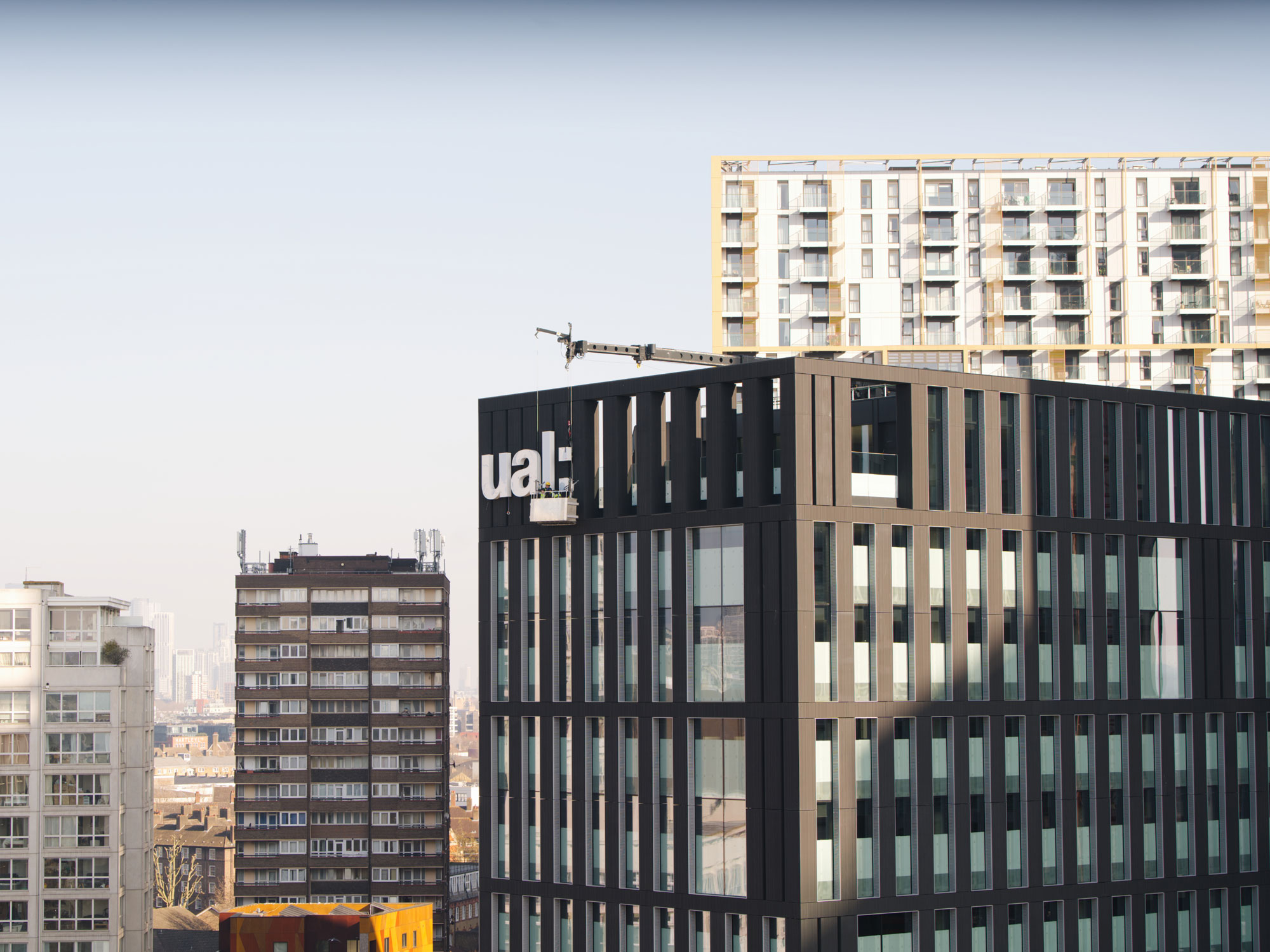 The UAL logo being installed by workers on a crane on the top of the new building for LCC - a large, black, broad tower block - on a blue sky day with other tall buildings in the background