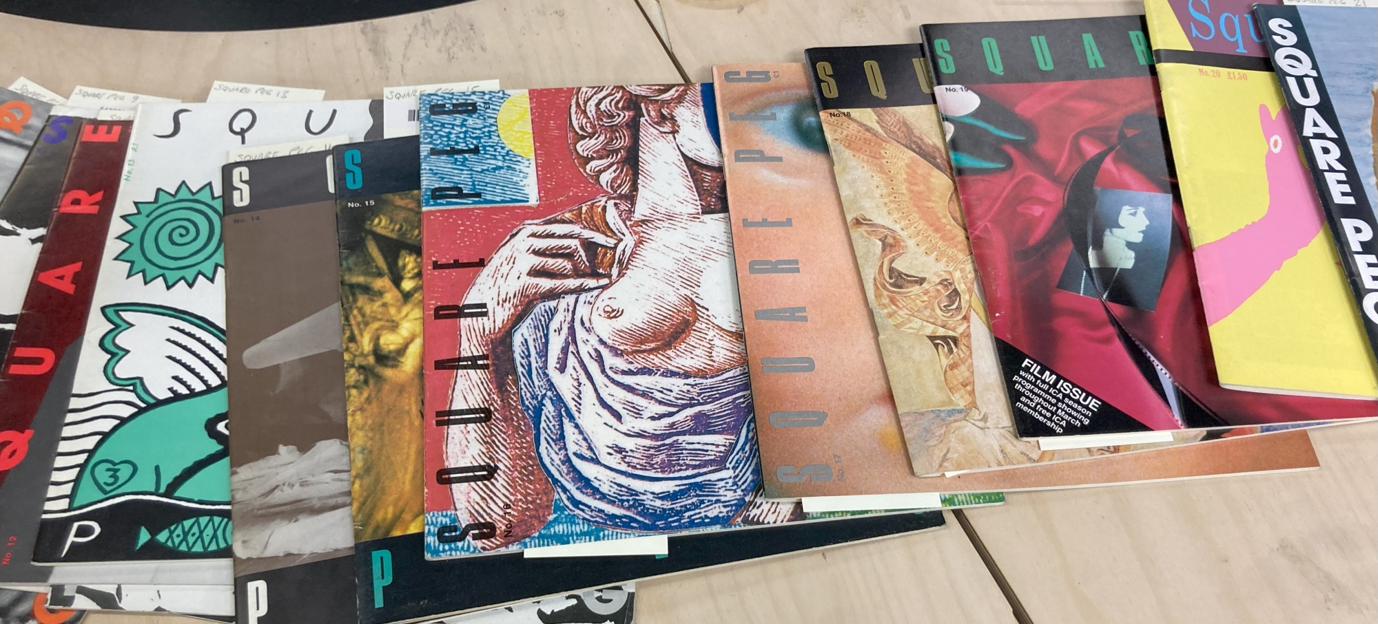 Colour photograph showing a spread of magazines titled Square Peg. The front of the magazines show a range of colourful cover artwork and photography. The cover artwork includes an art historical image of a figure wearing draped cloth with bare breasts. Another shows a photograph of a film strip with black and white portrait photography shown lying against a red satin background with green satin shoes in the top left corner.